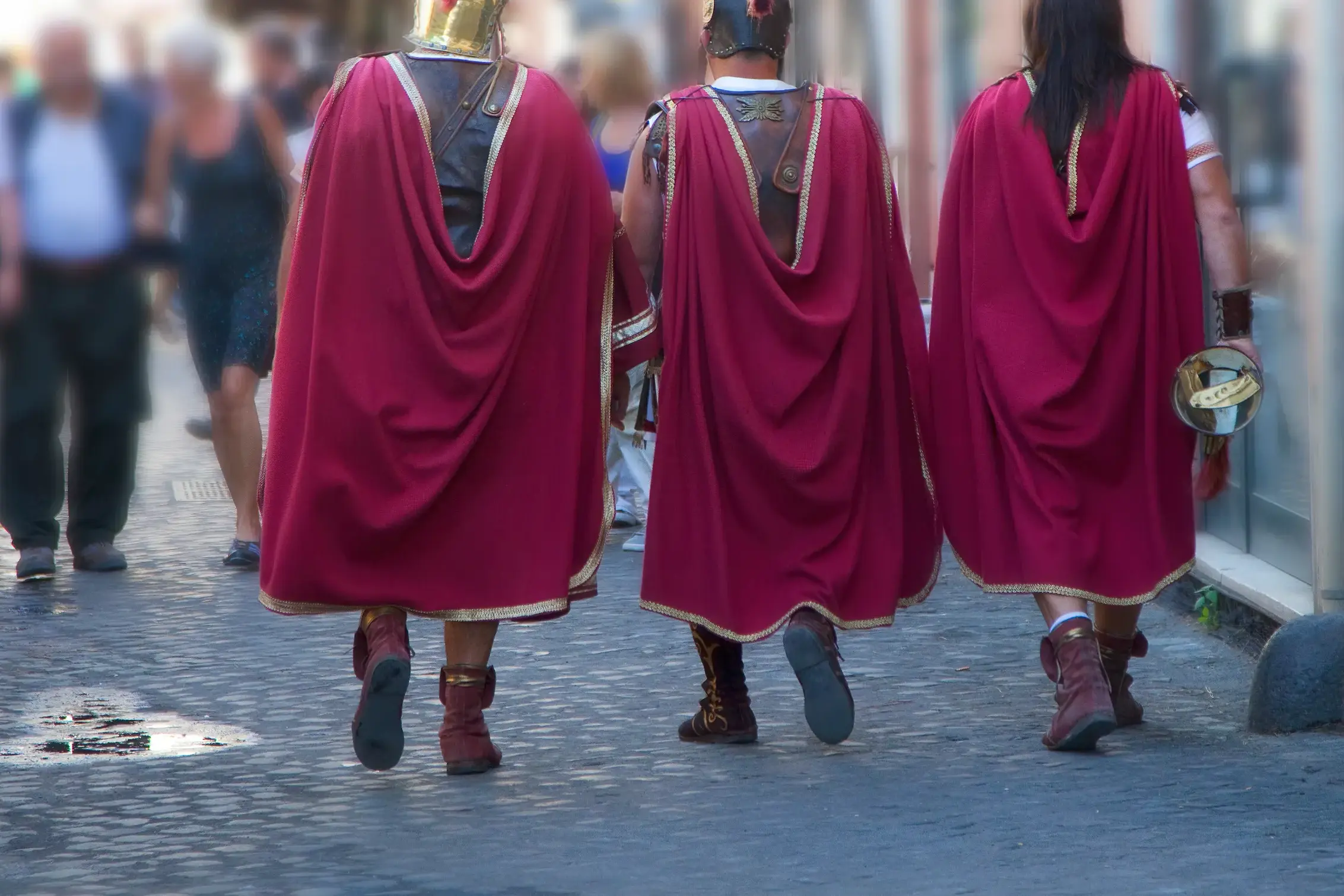 Drei als römische Gladiatoren verkleidete Männer mit roten Umhängen spazieren über das Kopfsteinpflaster einer Gasse in Rom.