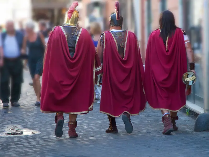 Drei als römische Gladiatoren verkleidete Männer mit roten Umhängen spazieren über das Kopfsteinpflaster einer Gasse in Rom.