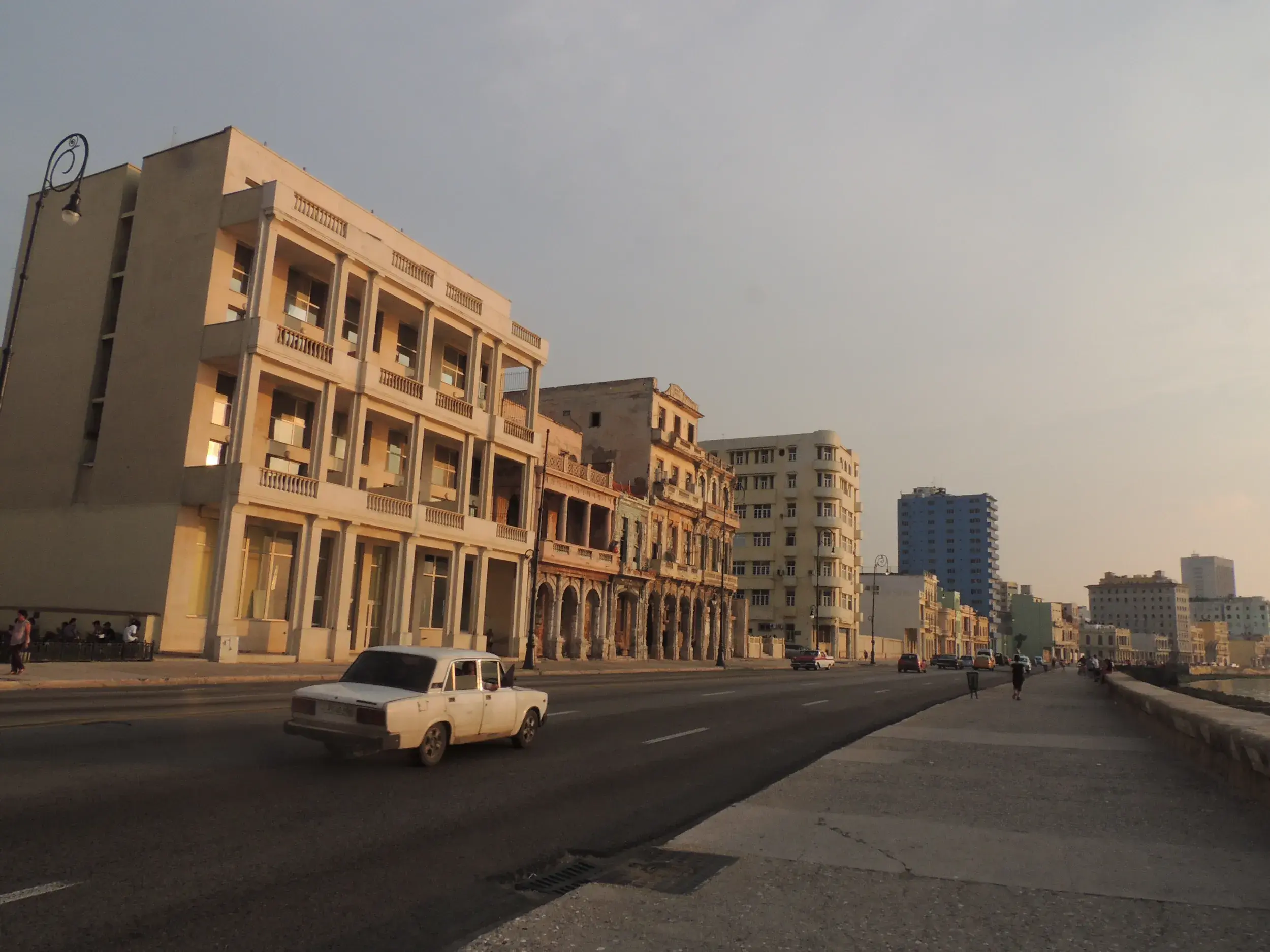 Ein weißer Oldtimer fährt auf der Uferpromenade Malecón in Havanna entlang historischer Kolonialbauten im Abendlicht.