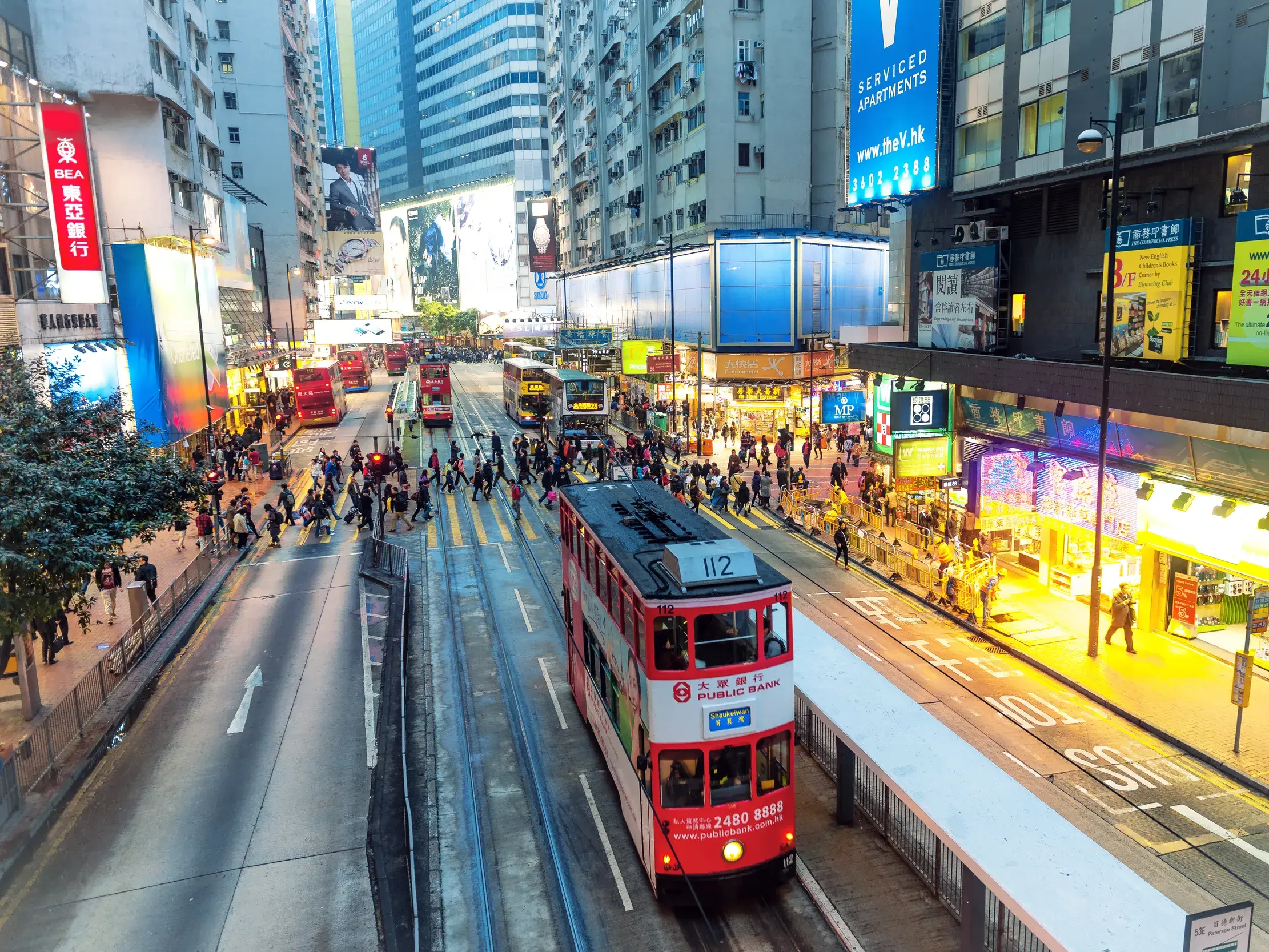 Rote Doppeldecker-Straßenbahn im dichten Abendverkehr einer belebten Hochhausschlucht in Hongkong.