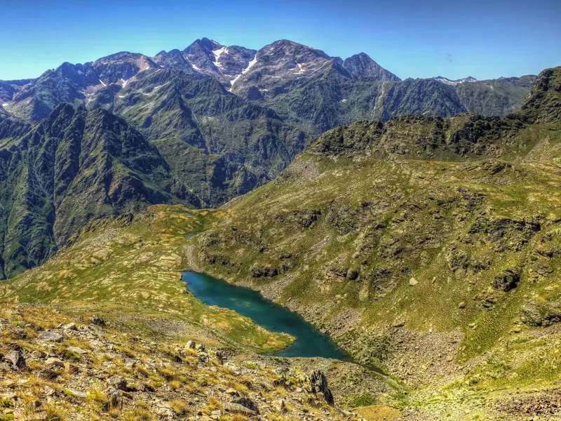 Blick von einem Wanderweg auf einen türkisen Karsee in den Pyrenäen, umgeben von grünen Hängen und schroffen Gipfeln Andorras.