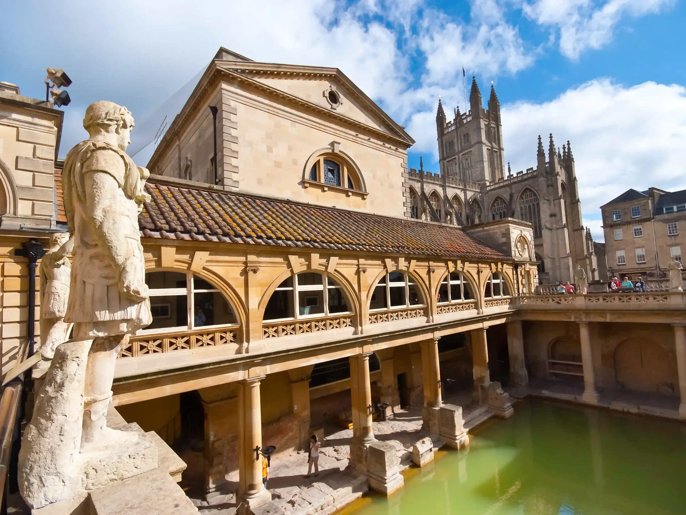 Römische Statue blickt auf das grüne Wasser des Großen Bades in Bath, im Hintergrund die Türme der Bath Abbey bei Sonnenschein.