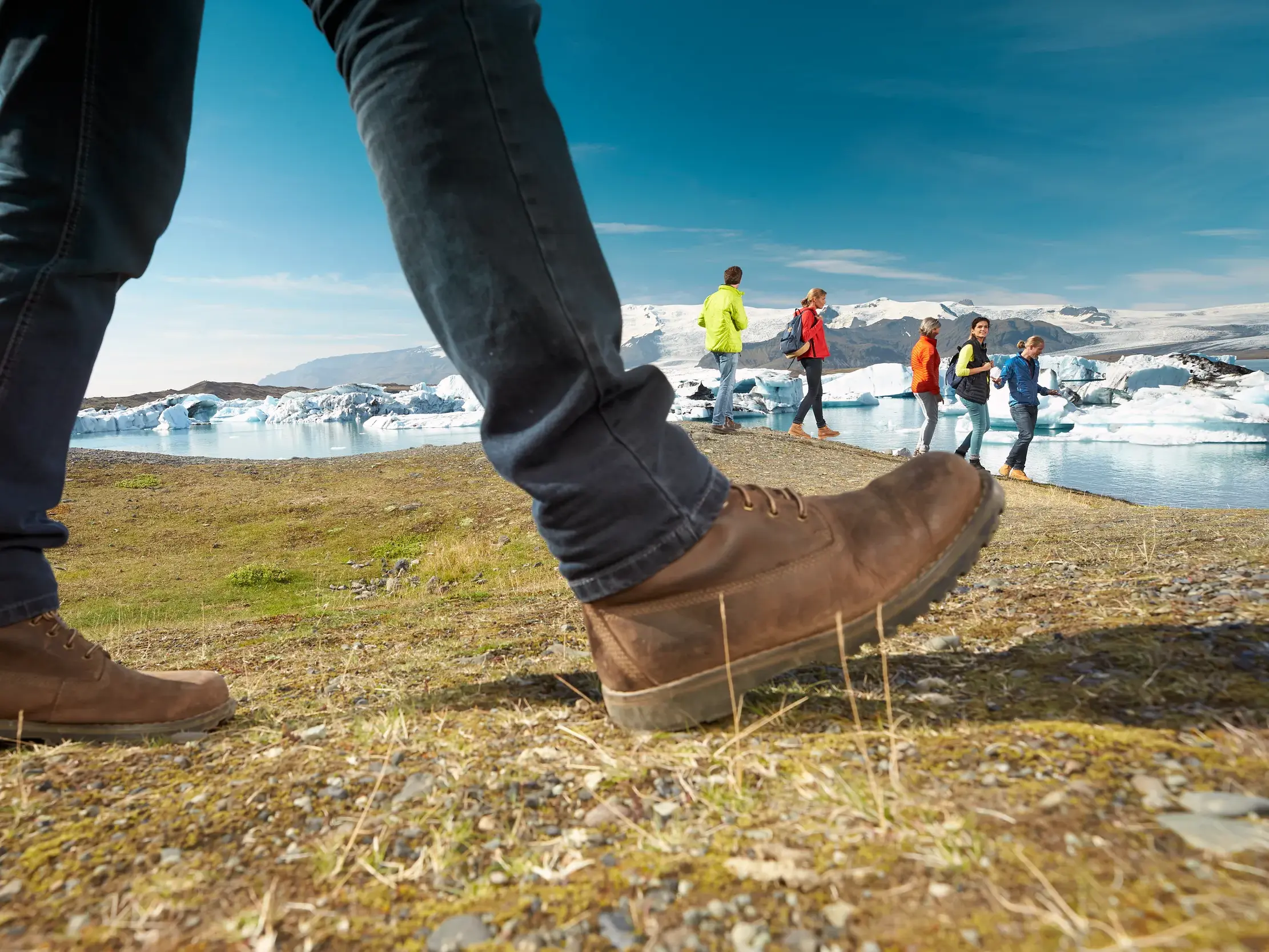 Wanderstiefel im Vordergrund bei einem Spaziergang am Ufer der Gletscherlagune Fjallsarlon mit treibenden Eisbergen in Island.