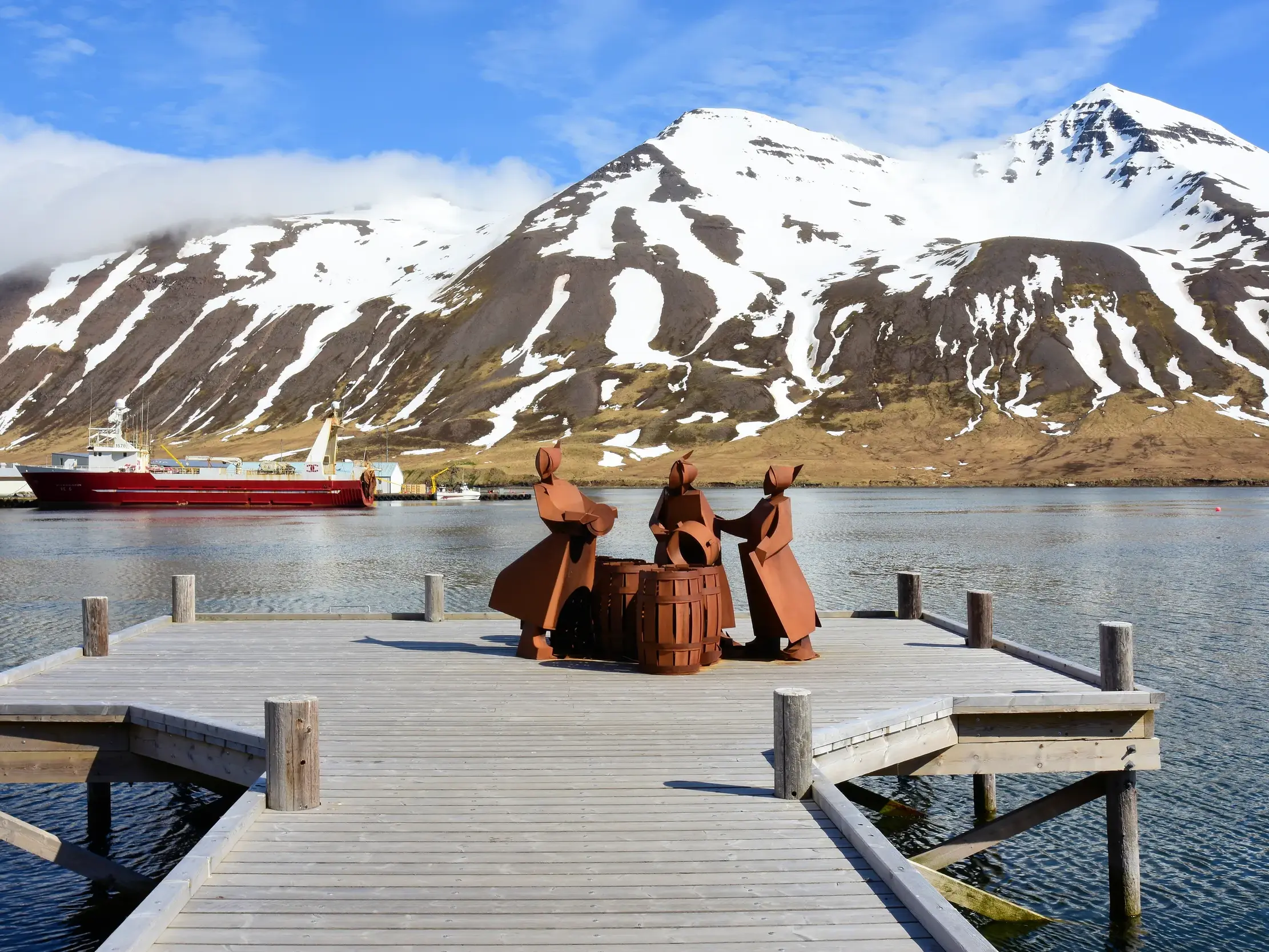 Denkmal für die Heringsfischerei auf einem Holzsteg im Fjord von Siglufjördur mit noch im Frühsommer schneebedeckten Bergen im Hintergrund.