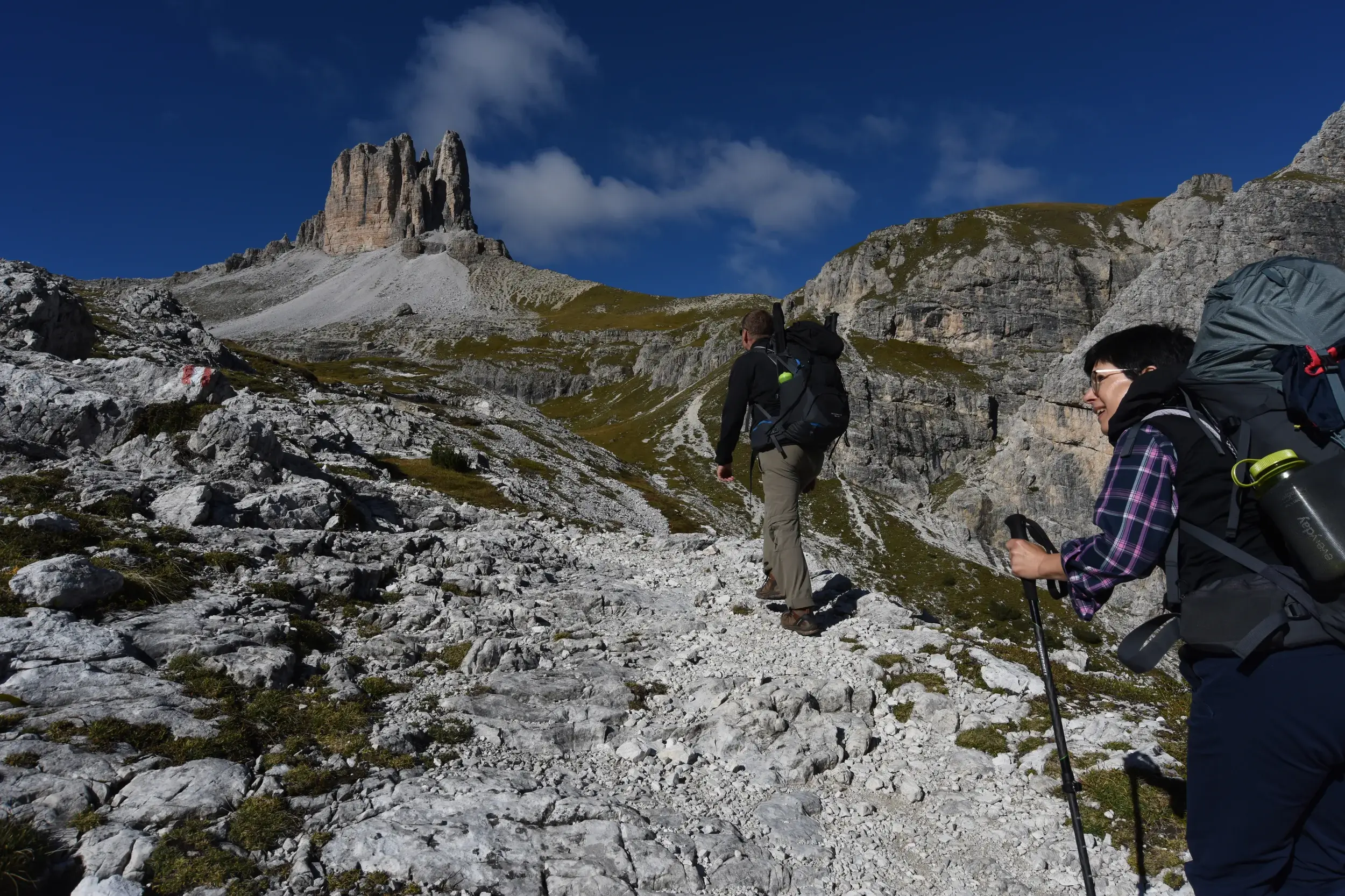 Zwei Wanderer mit Trekkingrucksäcken steigen auf einem steinigen Pfad auf, im Hintergrund der Toblinger Knoten in den Dolomiten.