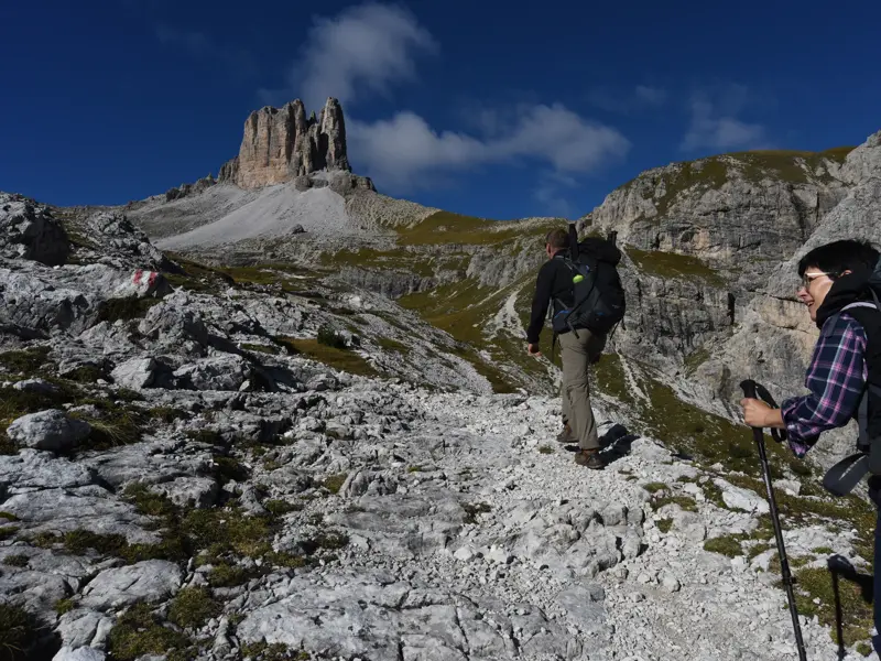 Zwei Wanderer mit Trekkingrucksäcken steigen auf einem steinigen Pfad auf, im Hintergrund der Toblinger Knoten in den Dolomiten.