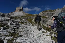 Zwei Wanderer mit Trekkingrucksäcken steigen auf einem steinigen Pfad auf, im Hintergrund der Toblinger Knoten in den Dolomiten.