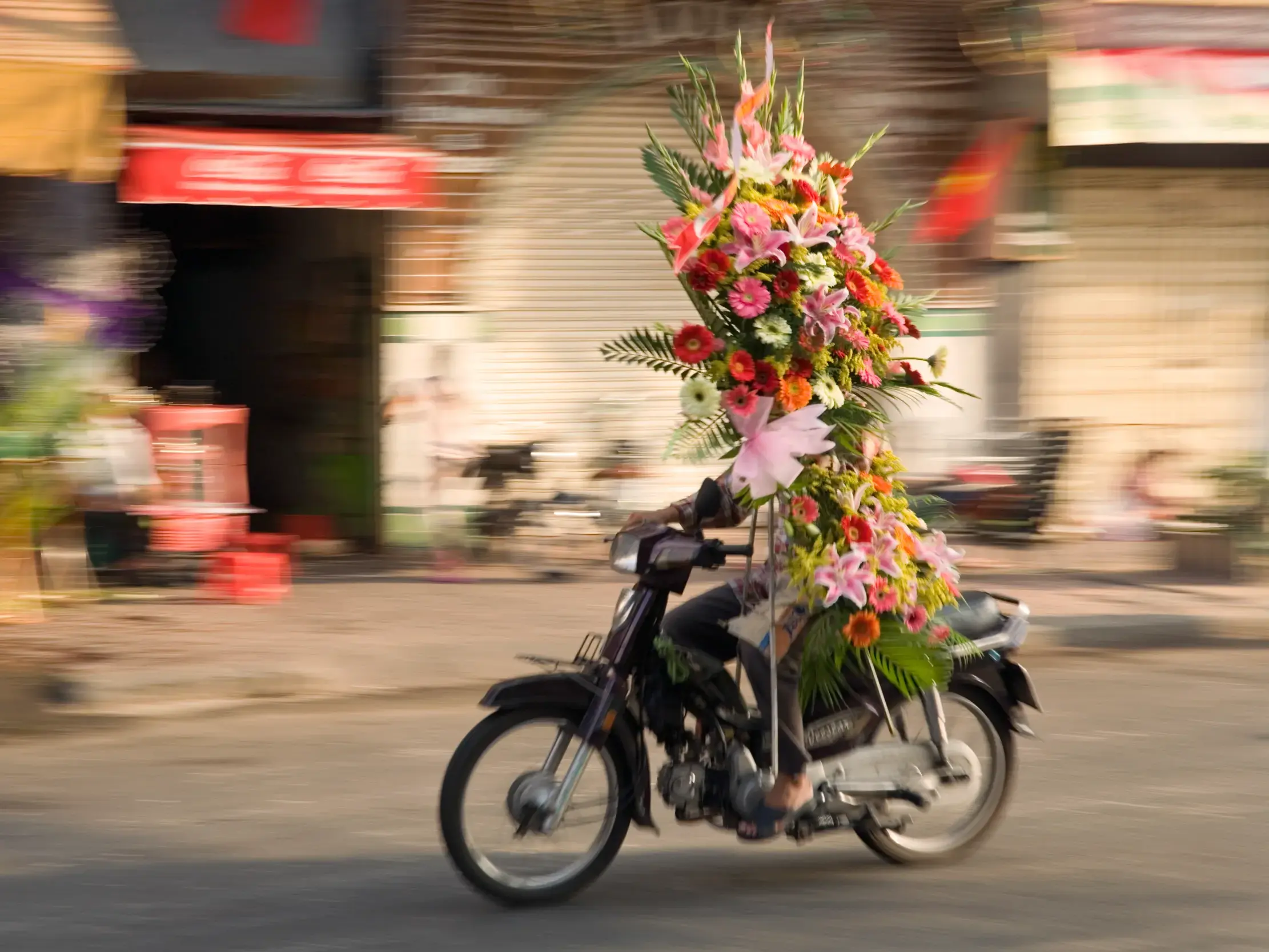 Ein Rollerfahrer transportiert ein turmhohes, farbenfrohes Blumengesteck durch den quirligen Straßenverkehr von Saigon, Vietnam.