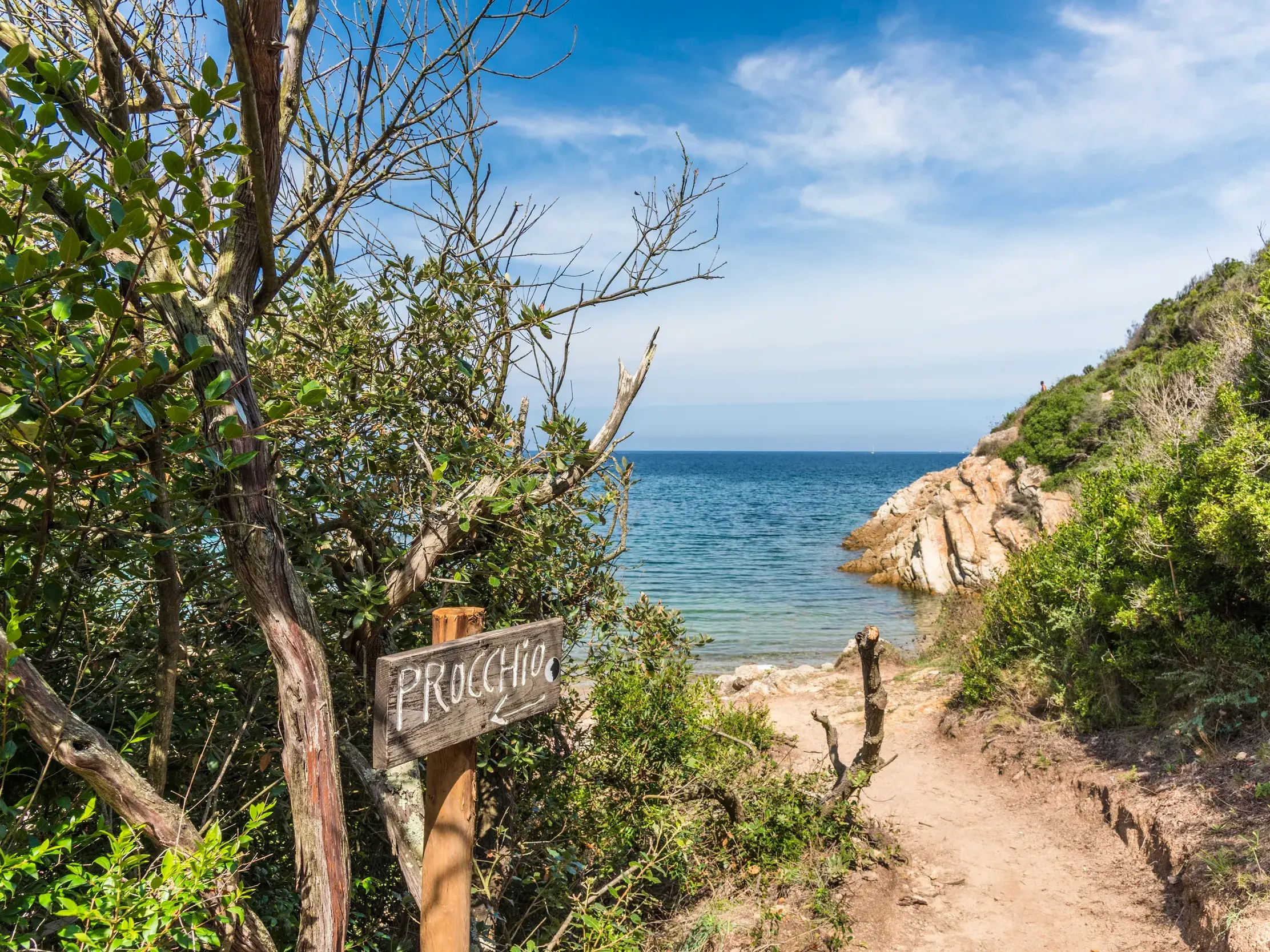 Wegweiser nach Procchio auf einem Küstenwanderweg auf Elba mit Blick auf eine malerische, felsige Bucht am Mittelmeer.