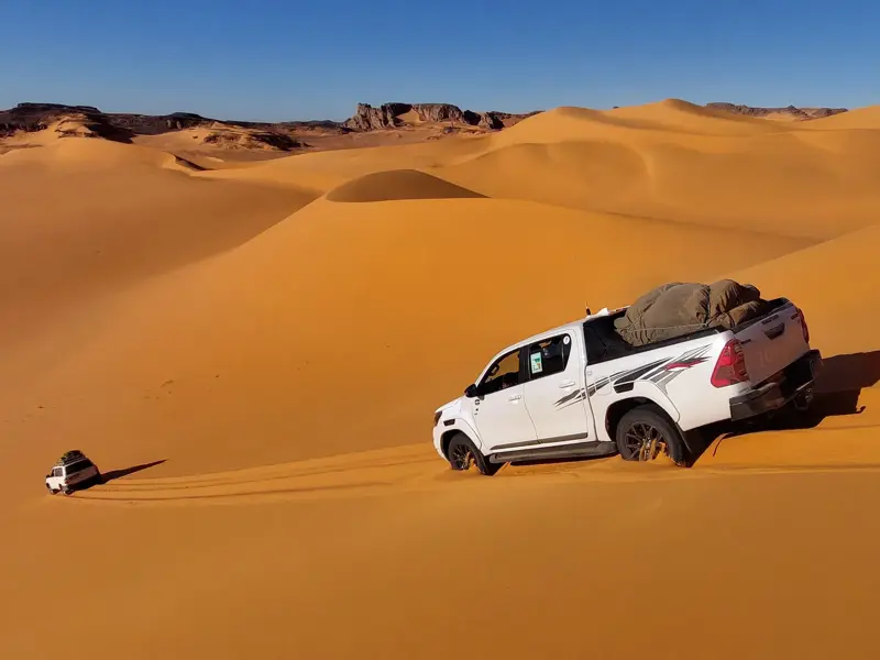 Zwei weiße Geländewagen bahnen sich ihren Weg durch die endlosen, orangefarbenen Sanddünen der Tadrart-Wüste in Algerien.