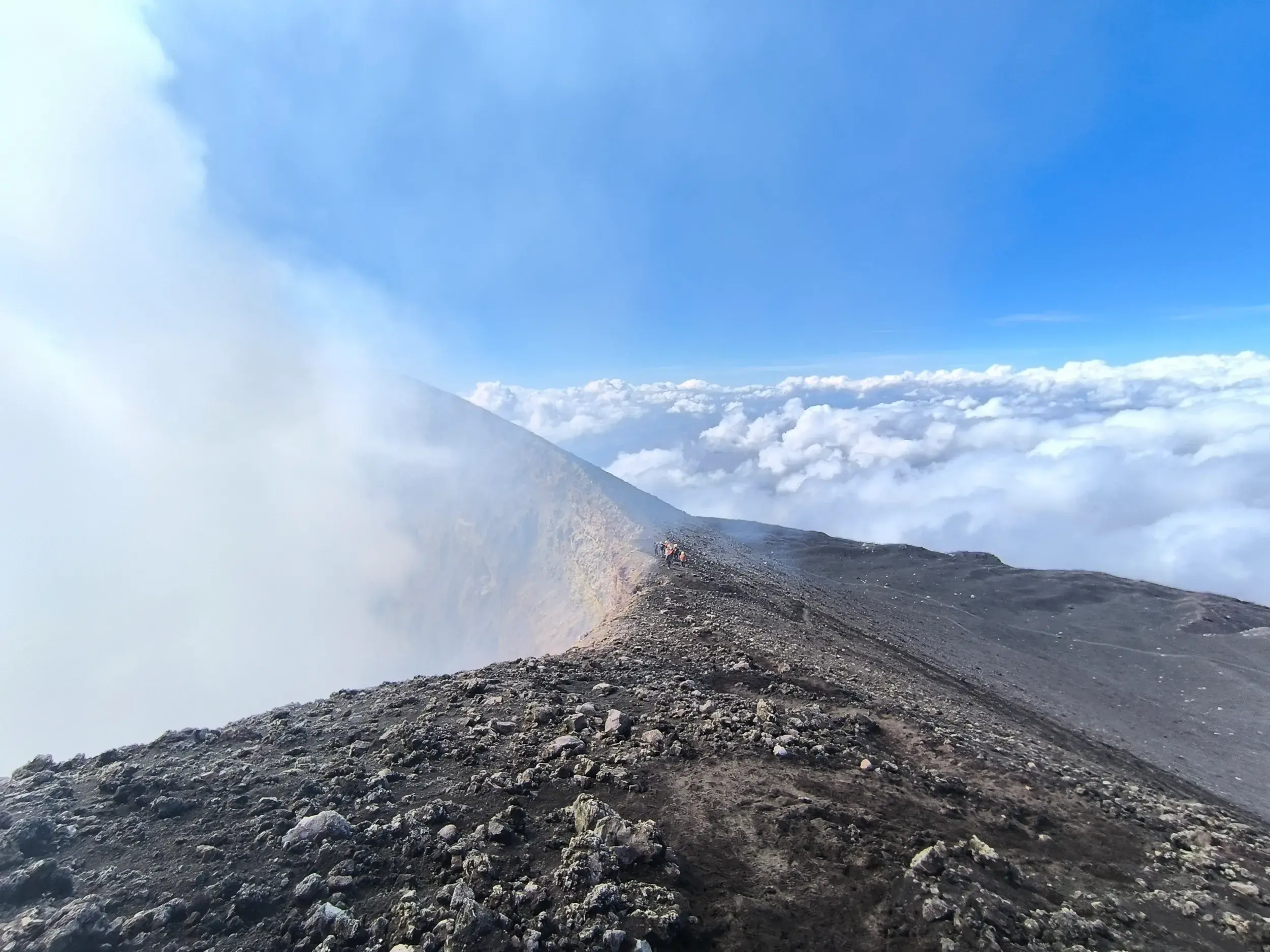 Eine Wandergruppe auf einem schmalen Grat am rauchenden Krater des Vulkans Ätna hoch über einem dichten Wolkenmeer.
