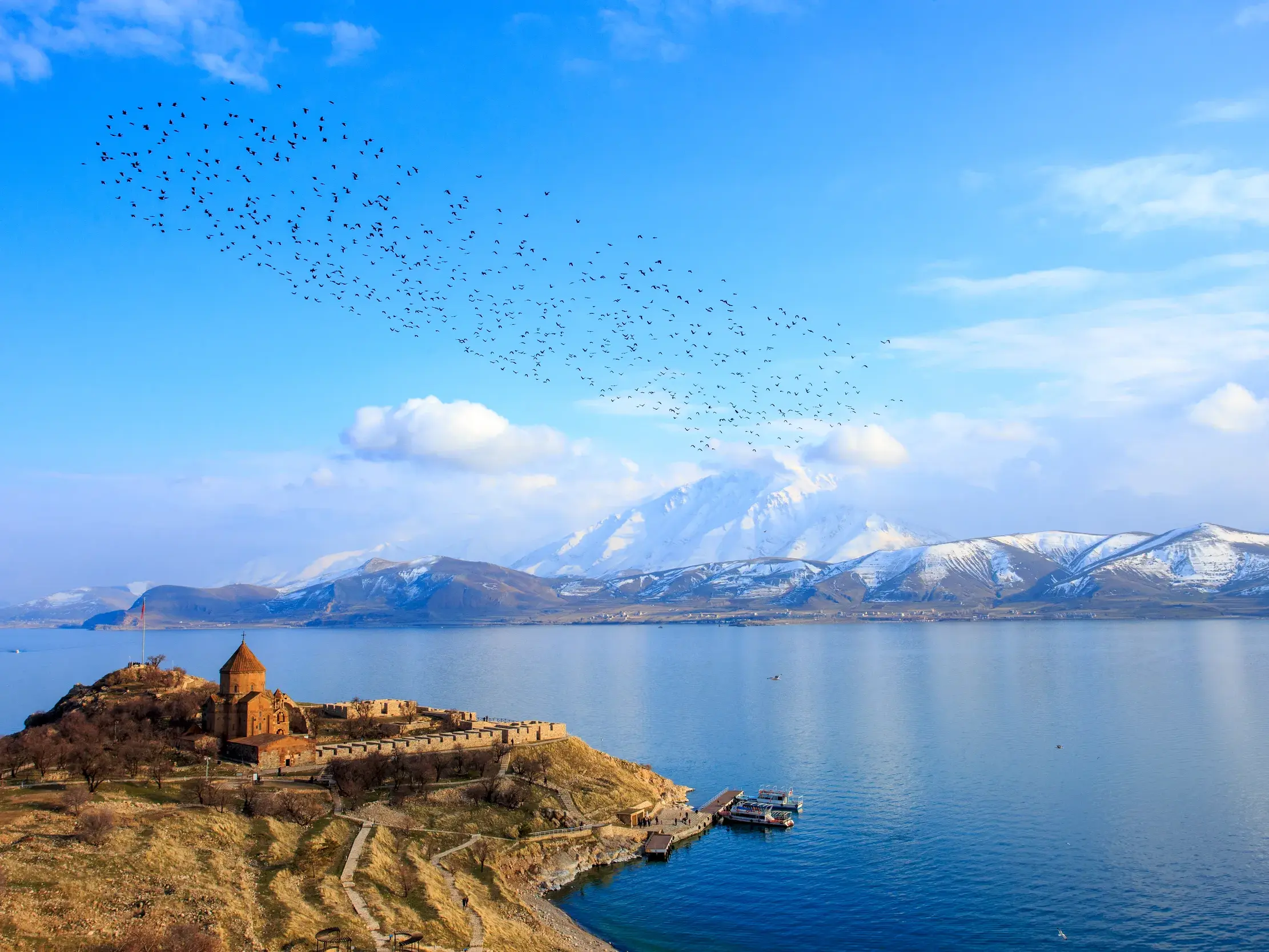 Rostbraunes Kloster Achtamar auf einer Insel im Vansee, im Hintergrund schneebedeckte Berge und ein Vogelschwarm am blauen Himmel.