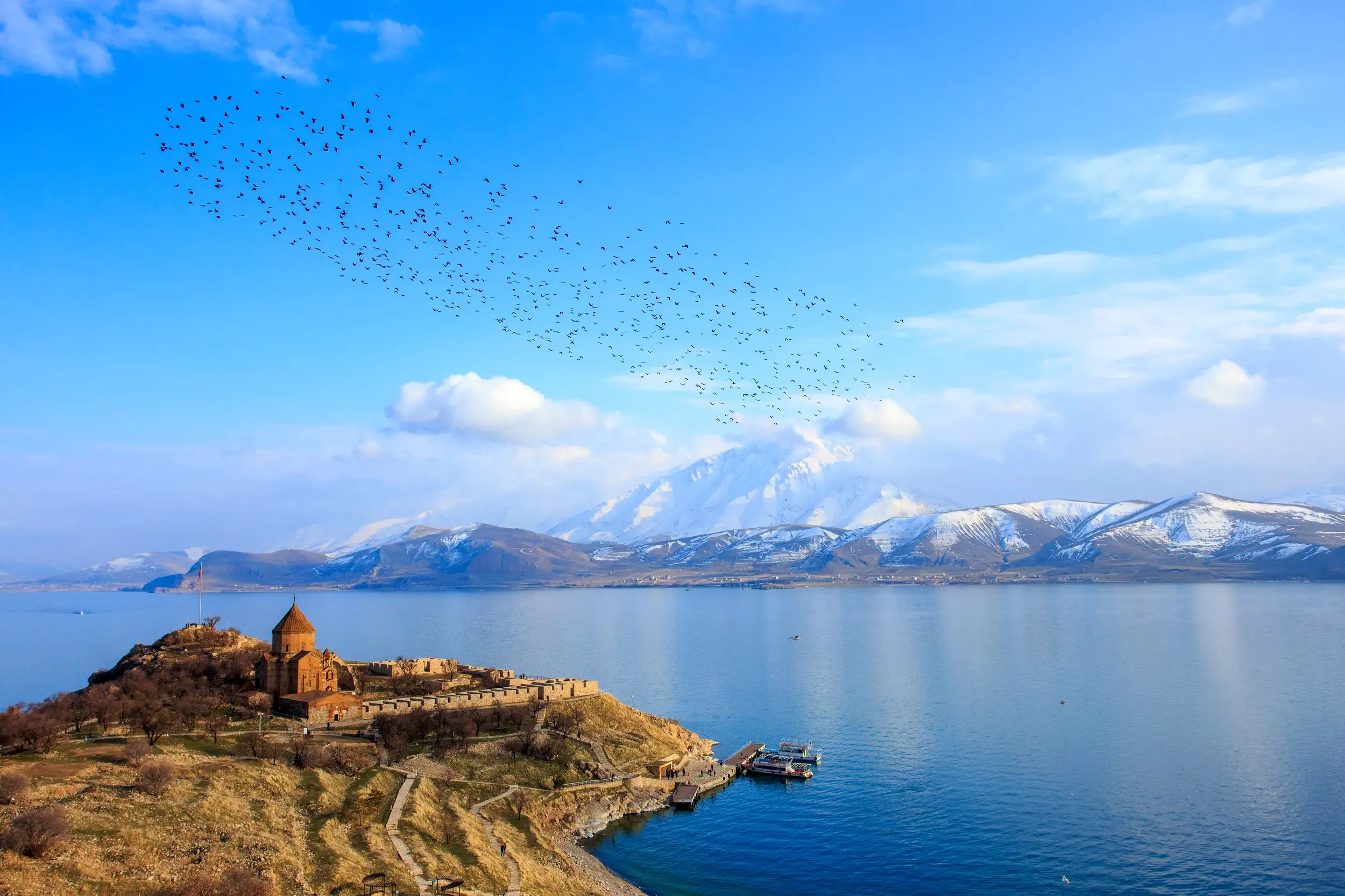 Rostbraunes Kloster Achtamar auf einer Insel im Vansee, im Hintergrund schneebedeckte Berge und ein Vogelschwarm am blauen Himmel.