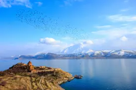 Rostbraunes Kloster Achtamar auf einer Insel im Vansee, im Hintergrund schneebedeckte Berge und ein Vogelschwarm am blauen Himmel.