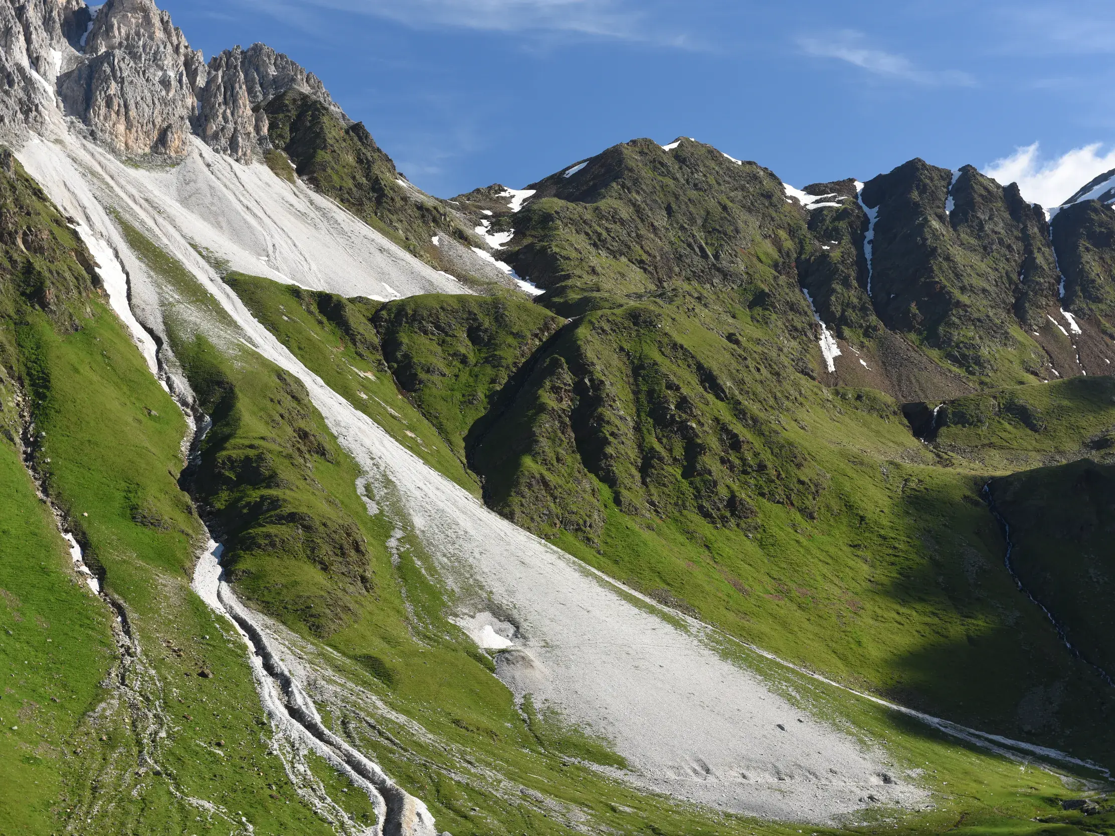 Schroffe Felsgipfel und weiße Geröllfelder in einer grünen alpinen Berglandschaft unter strahlend blauem Himmel.