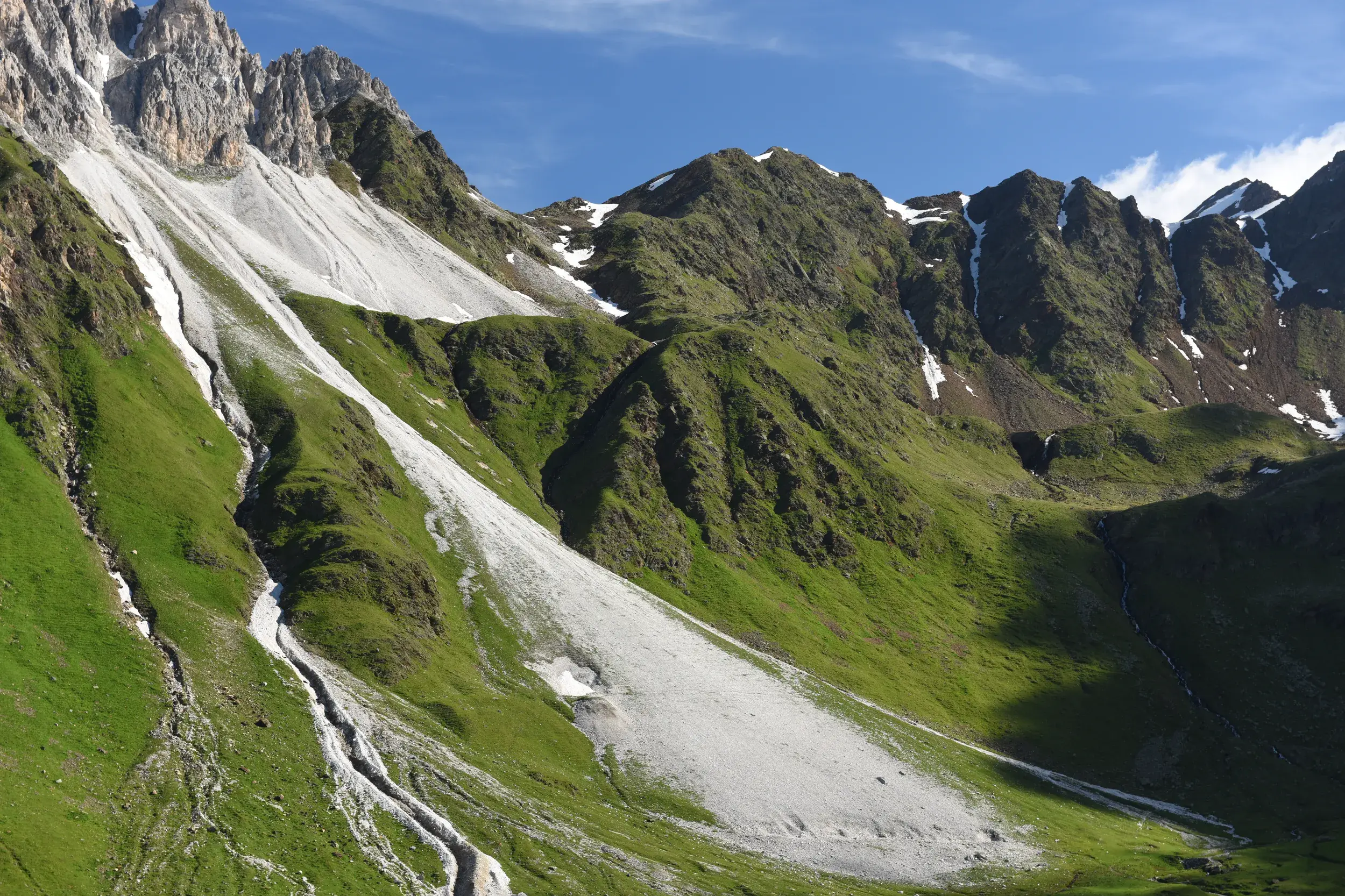 Schroffe Felsgipfel und weiße Geröllfelder in einer grünen alpinen Berglandschaft unter strahlend blauem Himmel.