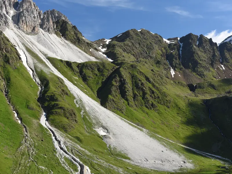 Schroffe Felsgipfel und weiße Geröllfelder in einer grünen alpinen Berglandschaft unter strahlend blauem Himmel.