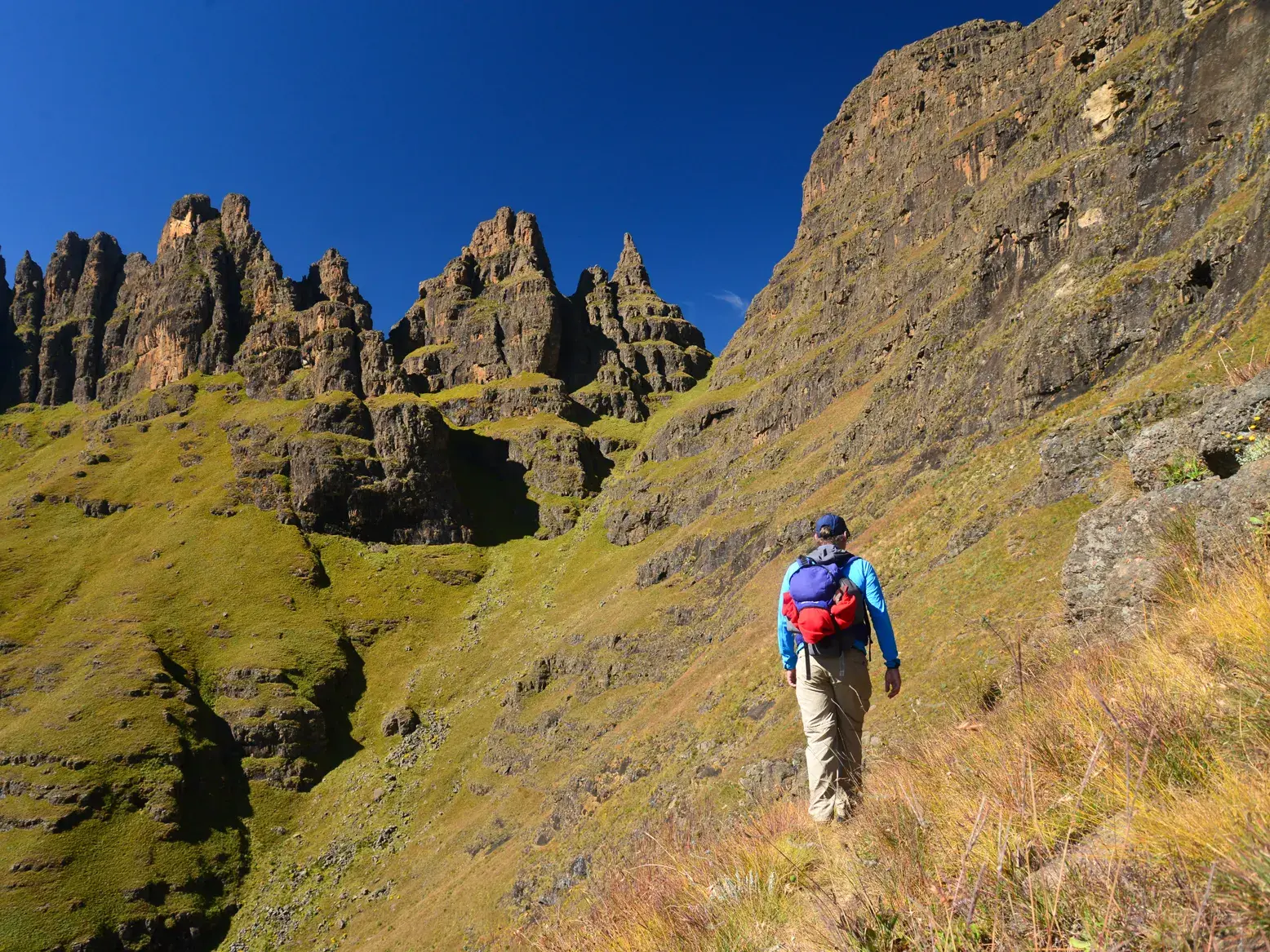 Ein Wanderer erklimmt einen grünen Hang beim Trekking in den zerklüfteten Drakensbergen Südafrikas unter klarem Himmel.