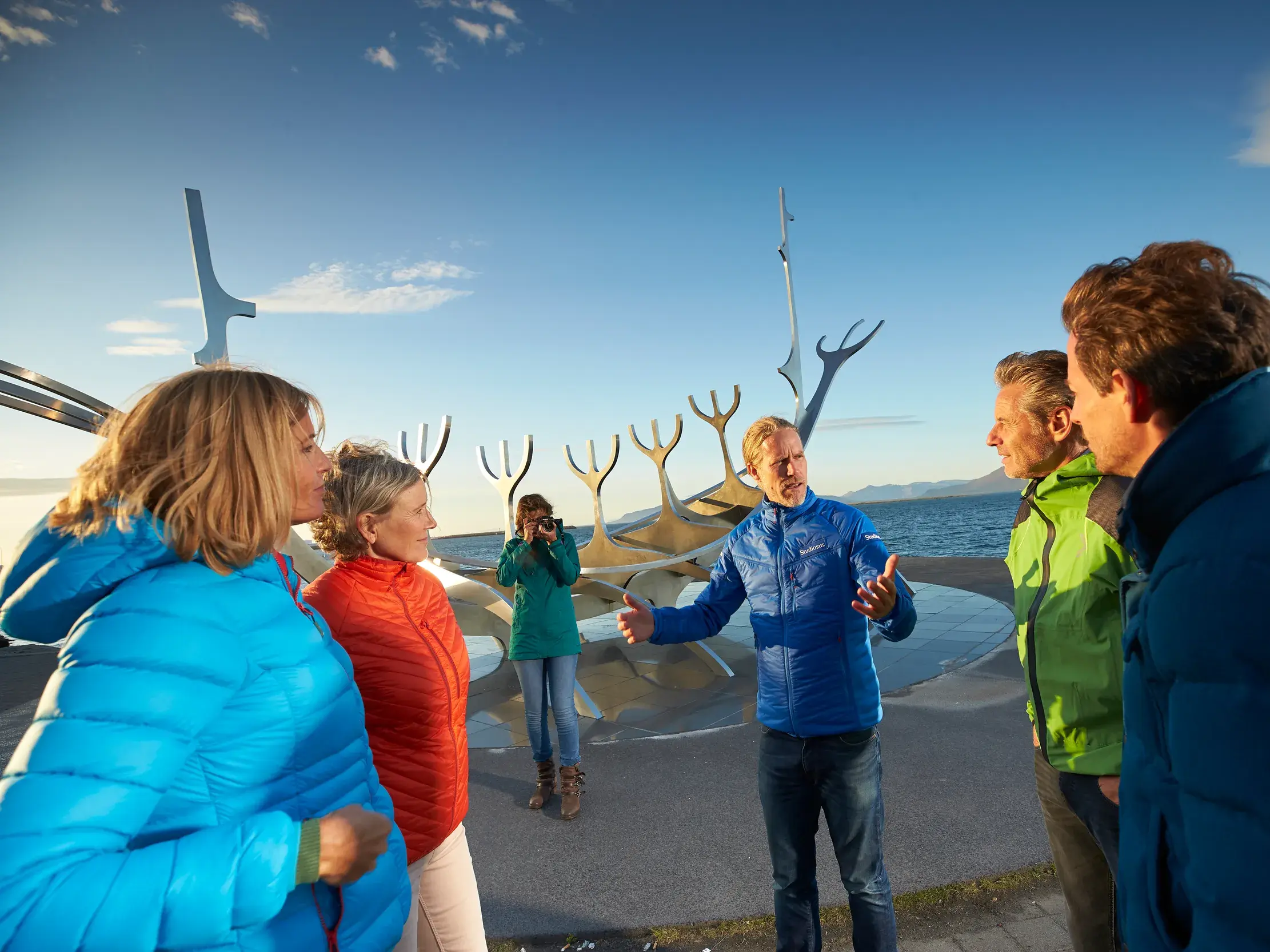 Ein Studiosus-Reiseleiter erklärt seiner Reisegruppe eine moderne Wikingerschiff-Skulptur am sonnigen Hafen von Reykjavik.