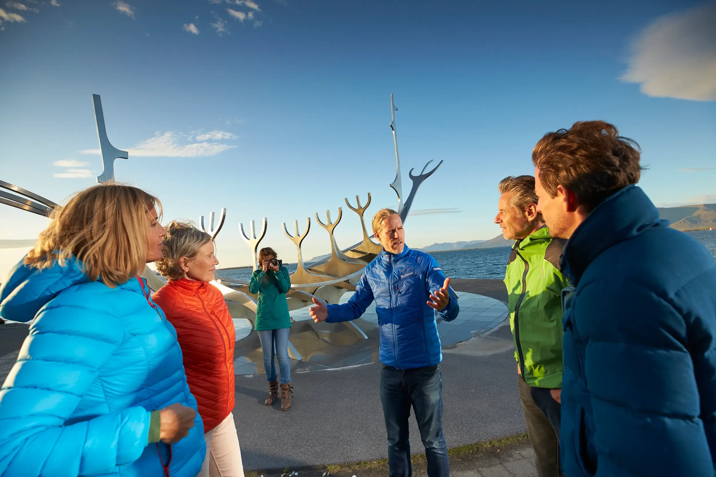 Ein Studiosus-Reiseleiter erklärt seiner Reisegruppe eine moderne Wikingerschiff-Skulptur am sonnigen Hafen von Reykjavik.