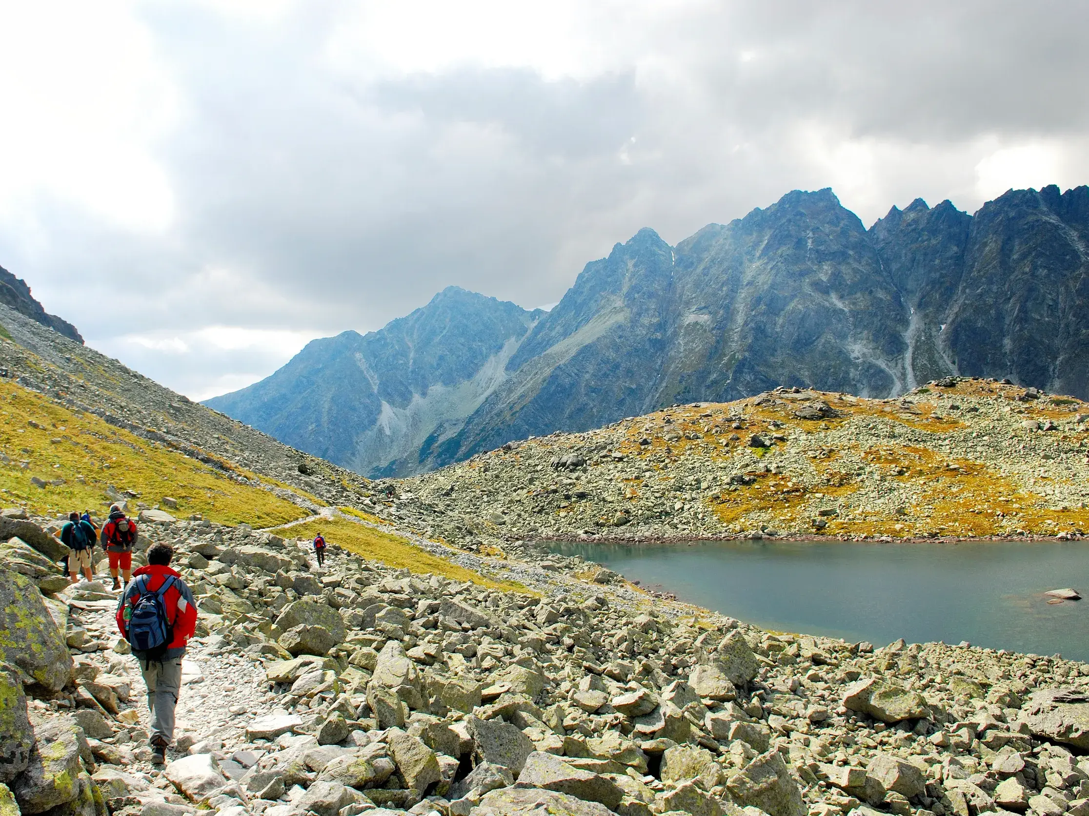 Eine Wandergruppe auf einem steinigen Pfad entlang eines Bergsees vor der Kulisse schroffer Gipfel in der Hohen Tatra.