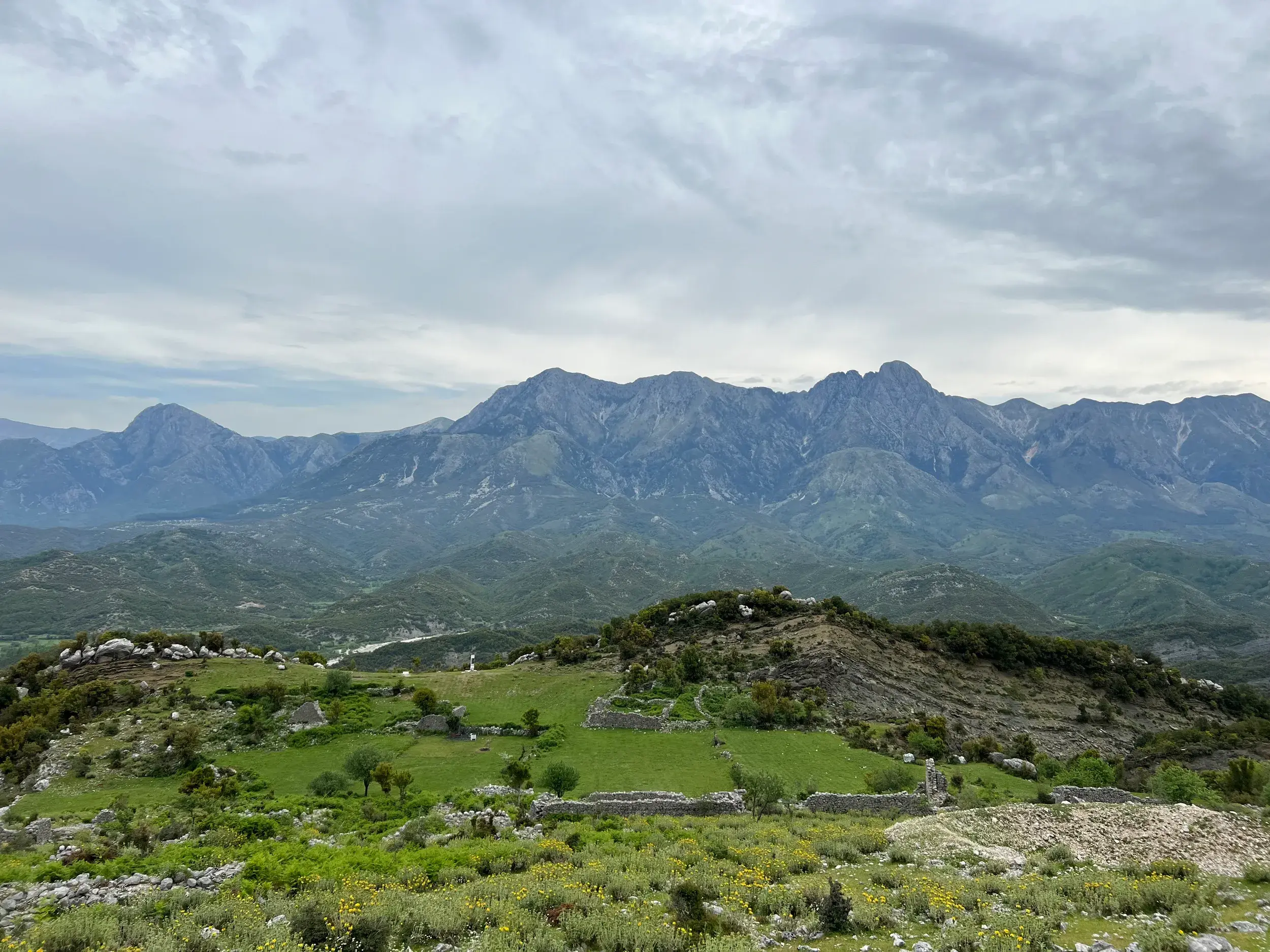 Panoramablick über grüne Hügel und Steinruinen auf die schroffe Bergkette des Ceraunischen Küstengebirges beim Trekking in Albanien.