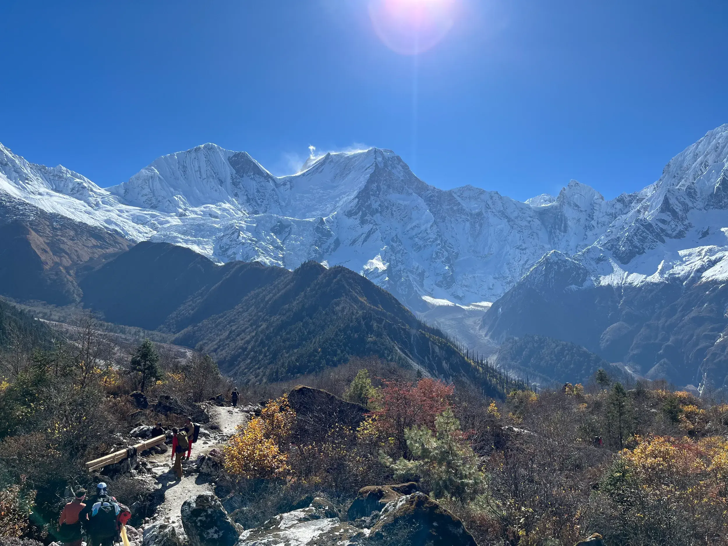 Wanderer und Träger auf einem Bergpfad vor der beeindruckenden Kulisse des schneebedeckten Manaslu-Himalaya bei Sonnenschein.