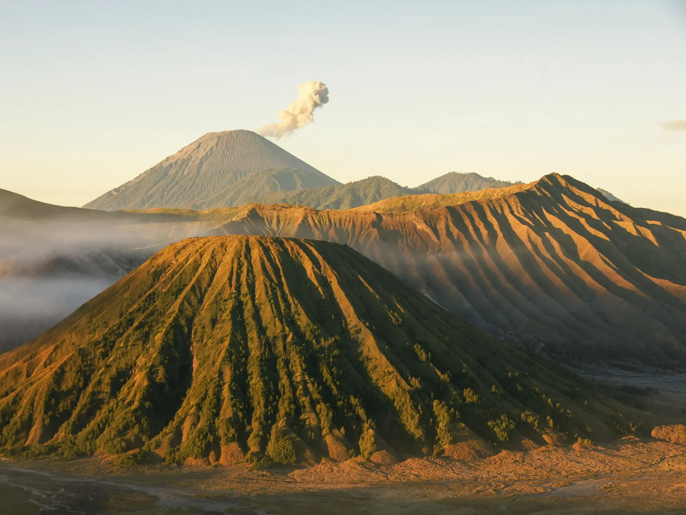 Die zerklüftete Vulkanlandschaft des Gunung Bromo in Java bei Sonnenaufgang, während im Hintergrund Rauch aufsteigt.