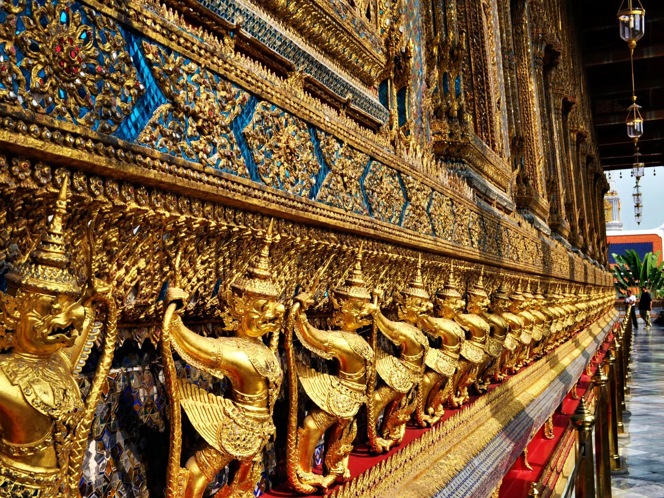 Goldene Garuda-Statuen stützen die kunstvoll mit blauen und goldenen Mosaiken verzierte Mauer des Großen Palastes in Bangkok.