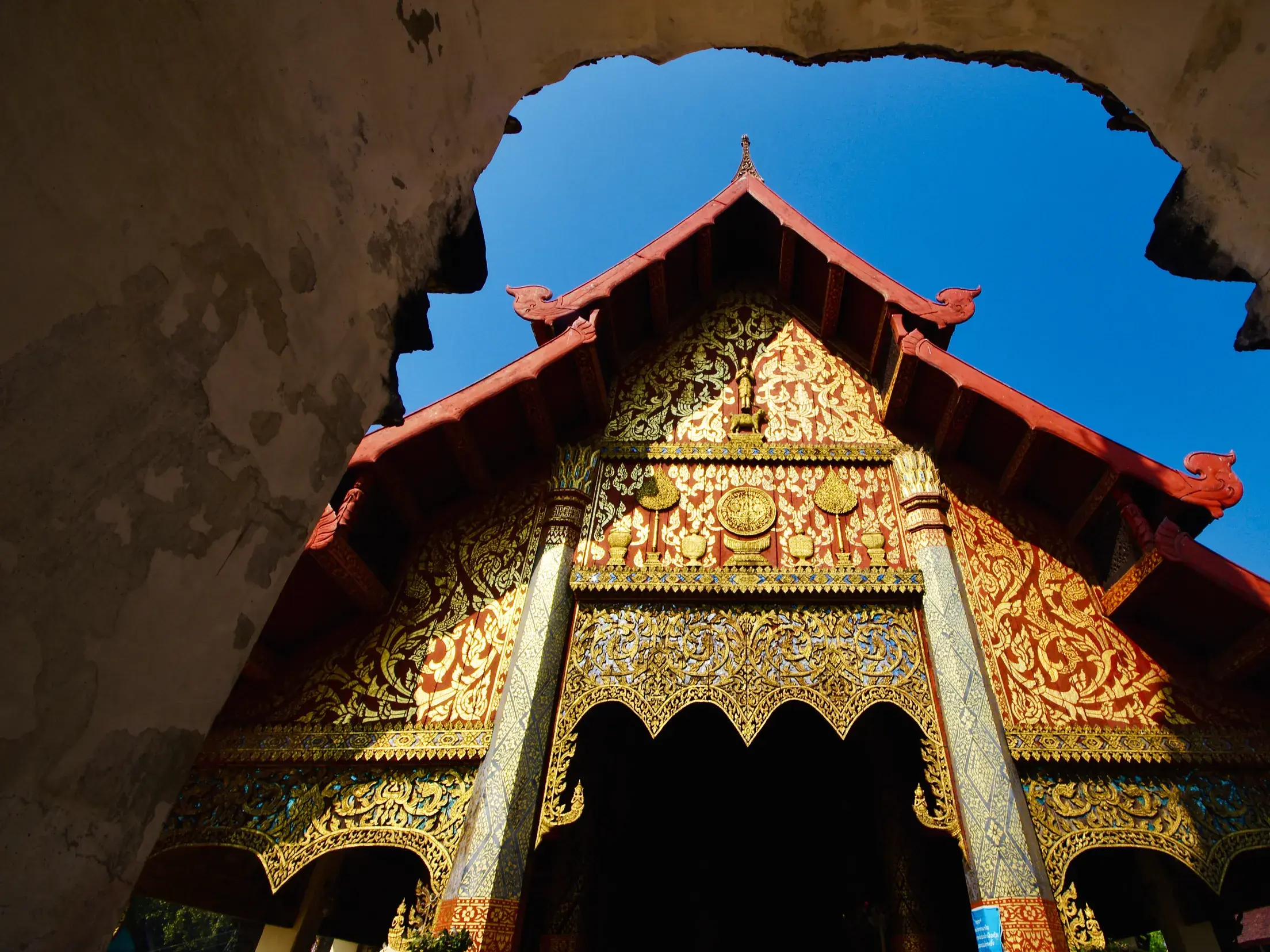 Blick durch ein steinernes Tor auf den reich verzierten Eingang des Tempels Wat Phra That Lampang Luang in Thailand.