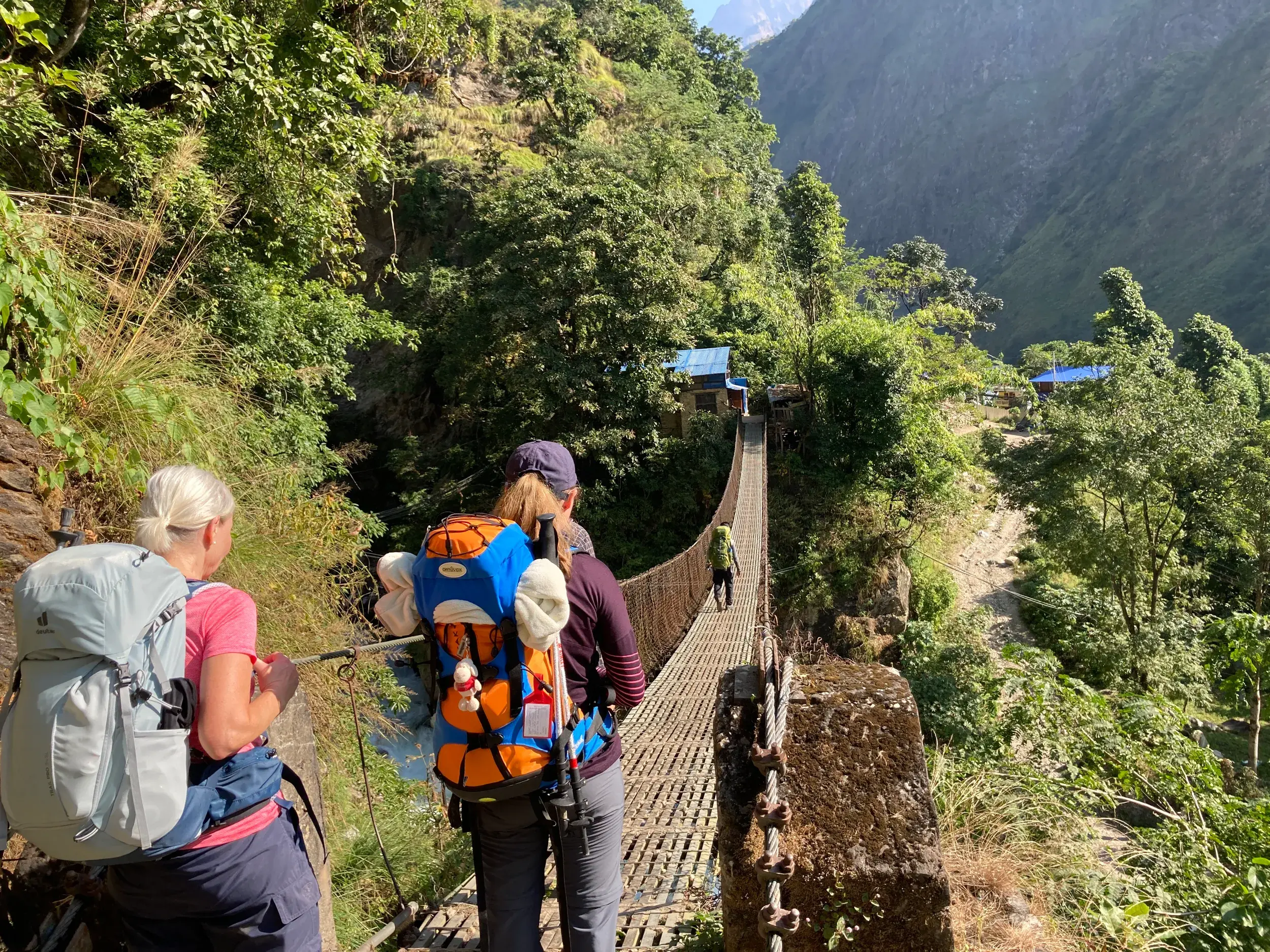 Eine Trekkinggruppe mit Rucksäcken überquert eine lange Hängebrücke über die grüne Schlucht des Buri Gandaki in Nepal.
