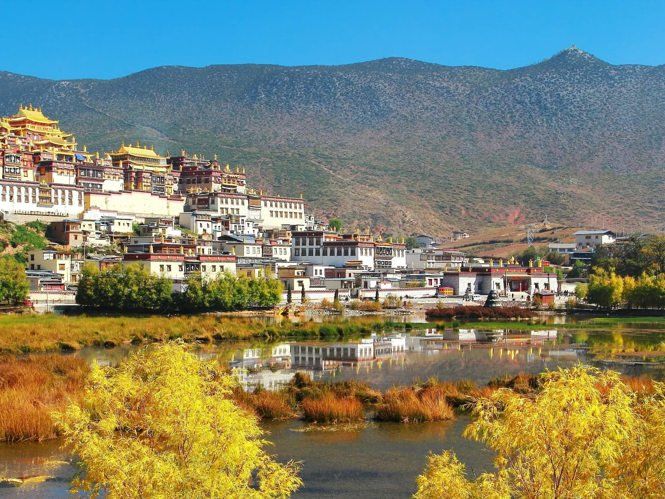 Das Ganden Songtsenling Kloster in Shangri-La spiegelt sich bei klarem Himmel in einem See vor einer Bergkette in Ost-Tibet.