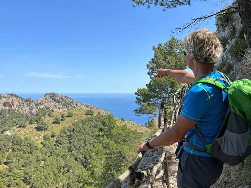 Wanderer mit Rucksack zeigt von einem Küstenpfad in der Serra de Tramuntana auf das weite, blaue Meer vor Mallorca.