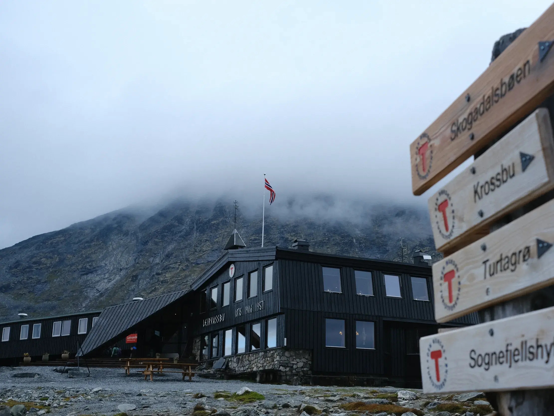 Hölzerner Wanderwegweiser vor der dunklen Leirvassbu Berghütte am Fuße eines nebligen Berges im Jotunheimen Nationalpark, Norwegen.