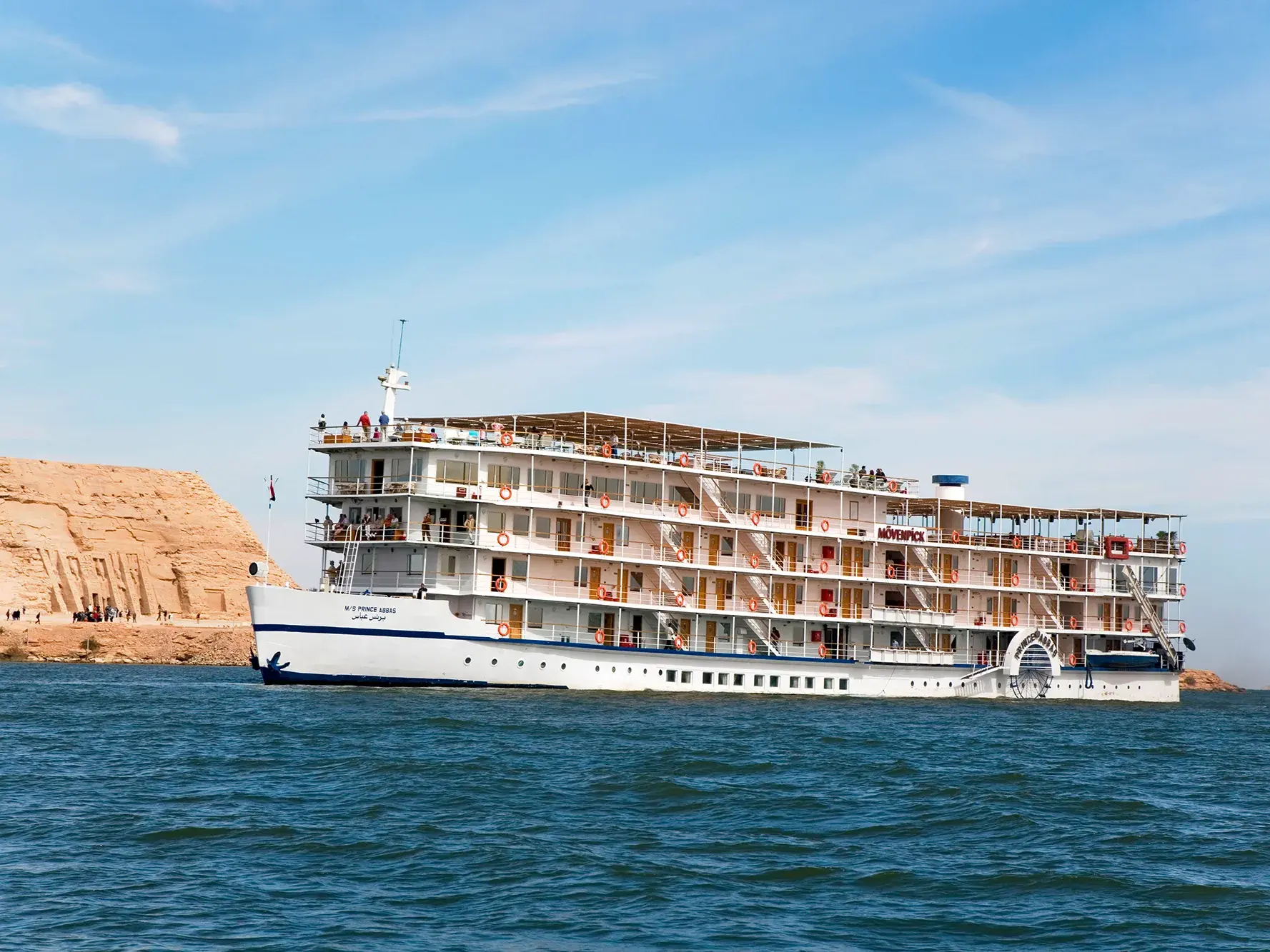 Das weiße Kreuzfahrtschiff MS Prince Abbas kreuzt bei Sonnenschein auf dem blauen Nassersee vor der Felsküste Ägyptens.