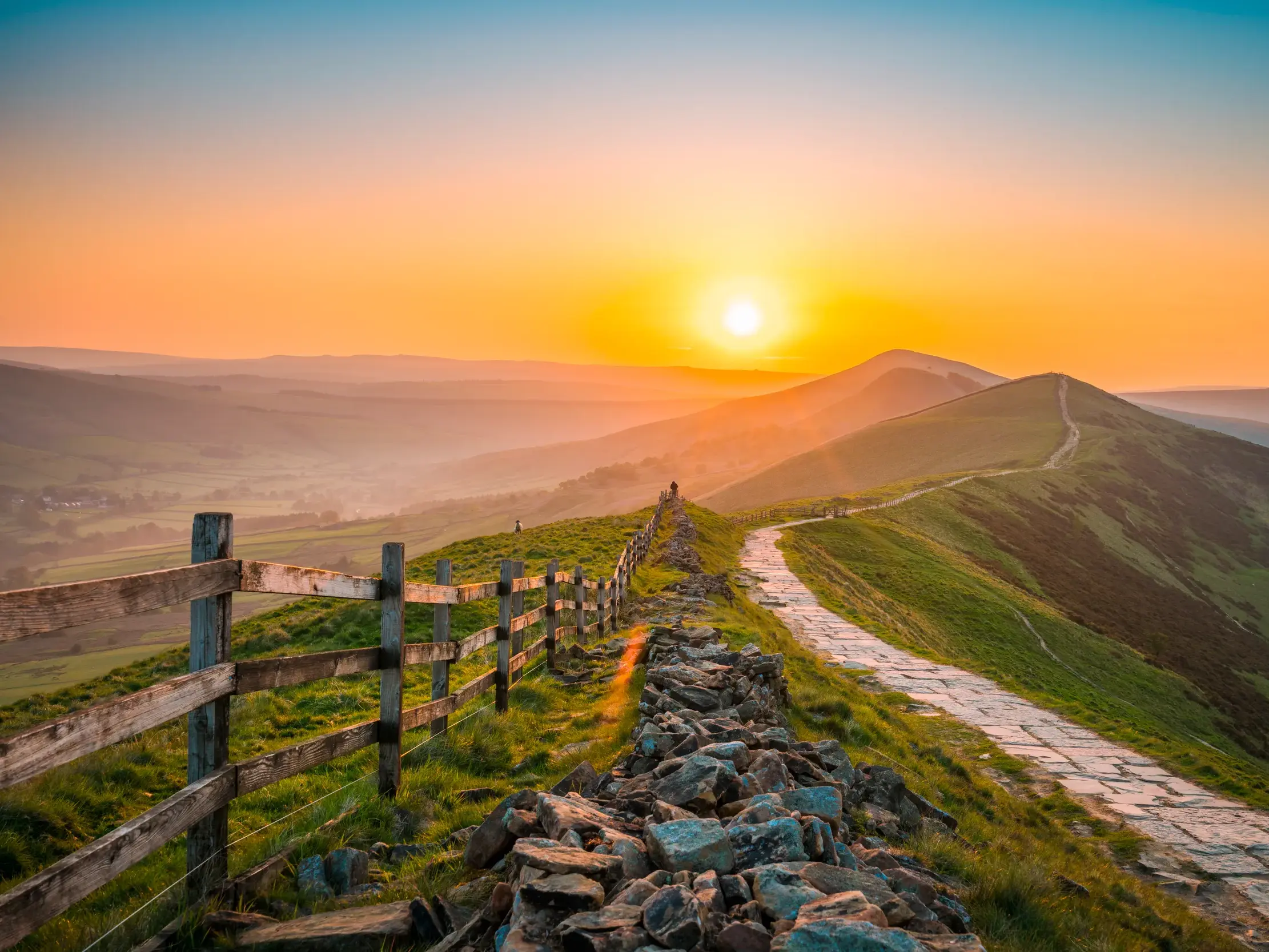 Ein gepflasterter Wanderweg führt bei Sonnenuntergang entlang eines Grats durch die grünen Hügel des Peak District.