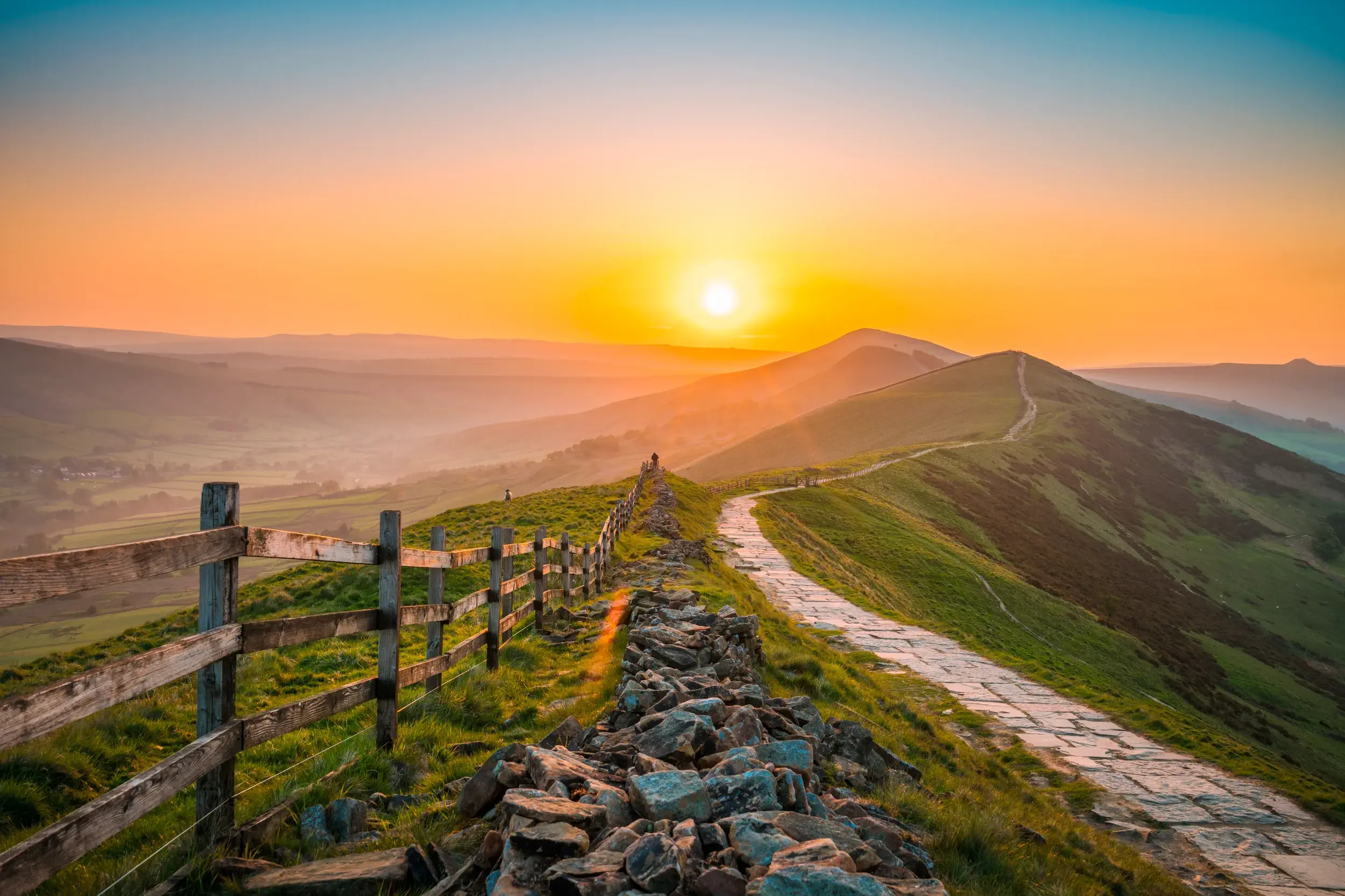 Ein gepflasterter Wanderweg führt bei Sonnenuntergang entlang eines Grats durch die grünen Hügel des Peak District.