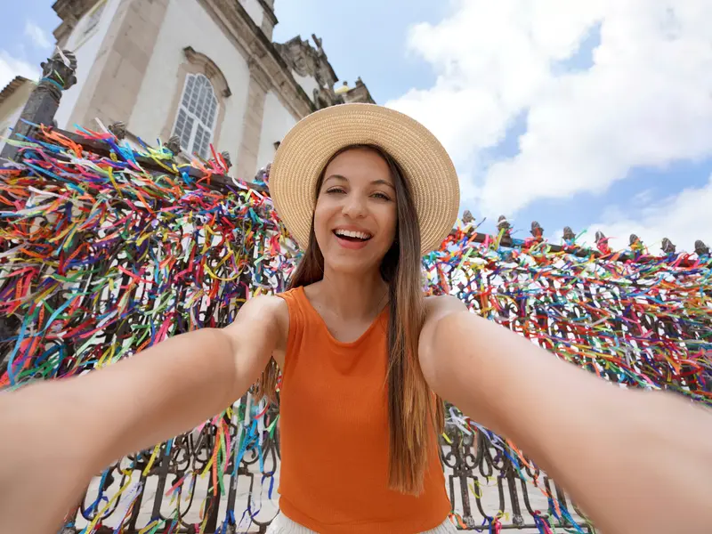 Lachende junge Frau mit Strohhut macht ein Selfie vor den bunten Wunschbändern der berühmten Bonfim-Kirche in Salvador.