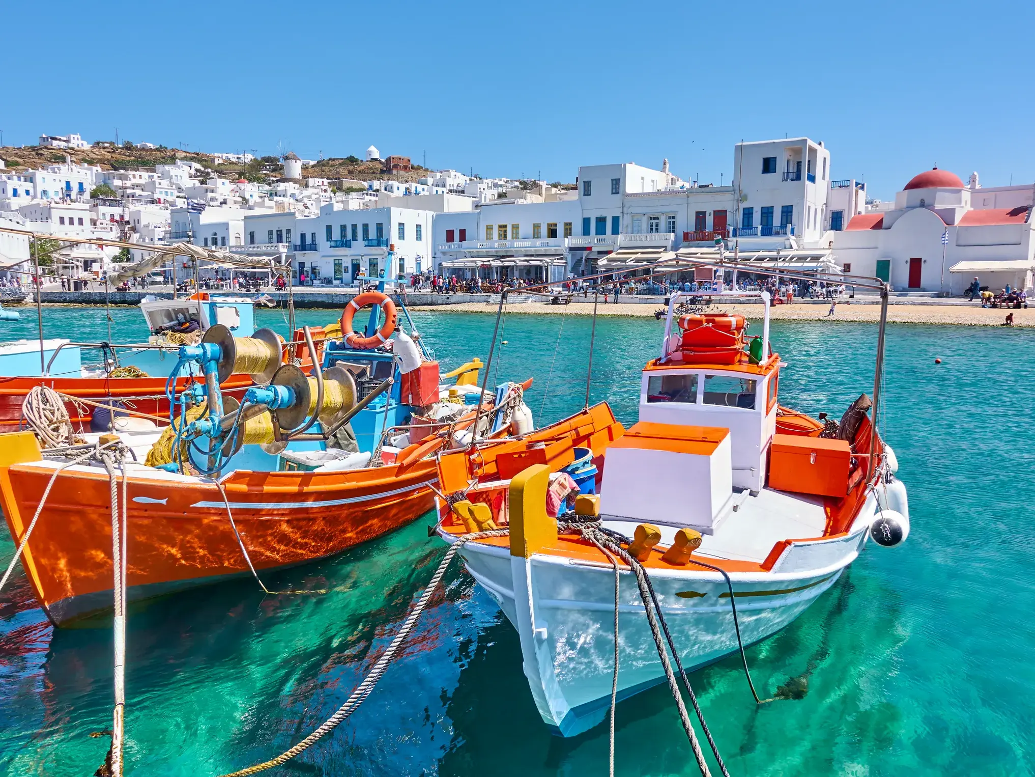 Bunte Fischerboote im türkisfarbenen Wasser des Hafens von Mykonos mit Blick auf die weißen Häuser und Windmühlen der Kykladeninsel.