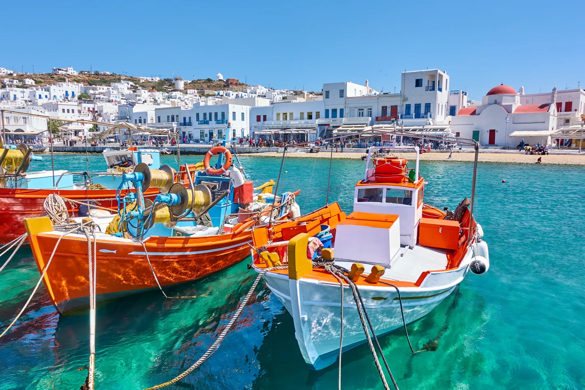Bunte Fischerboote im türkisfarbenen Wasser des Hafens von Mykonos mit Blick auf die weißen Häuser und Windmühlen der Kykladeninsel.