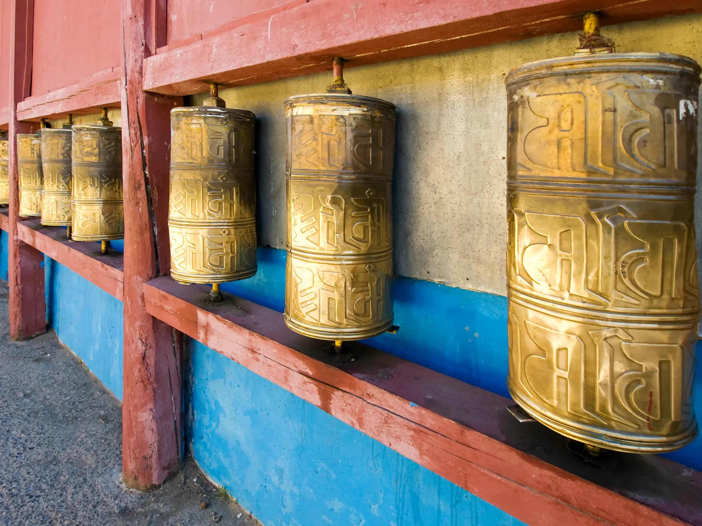 Eine Reihe goldener Gebetsmühlen mit eingravierten Glückssymbolen in einem rot-blauen Holzrahmen im Gandan-Kloster in Ulan Bator, Mongolei.