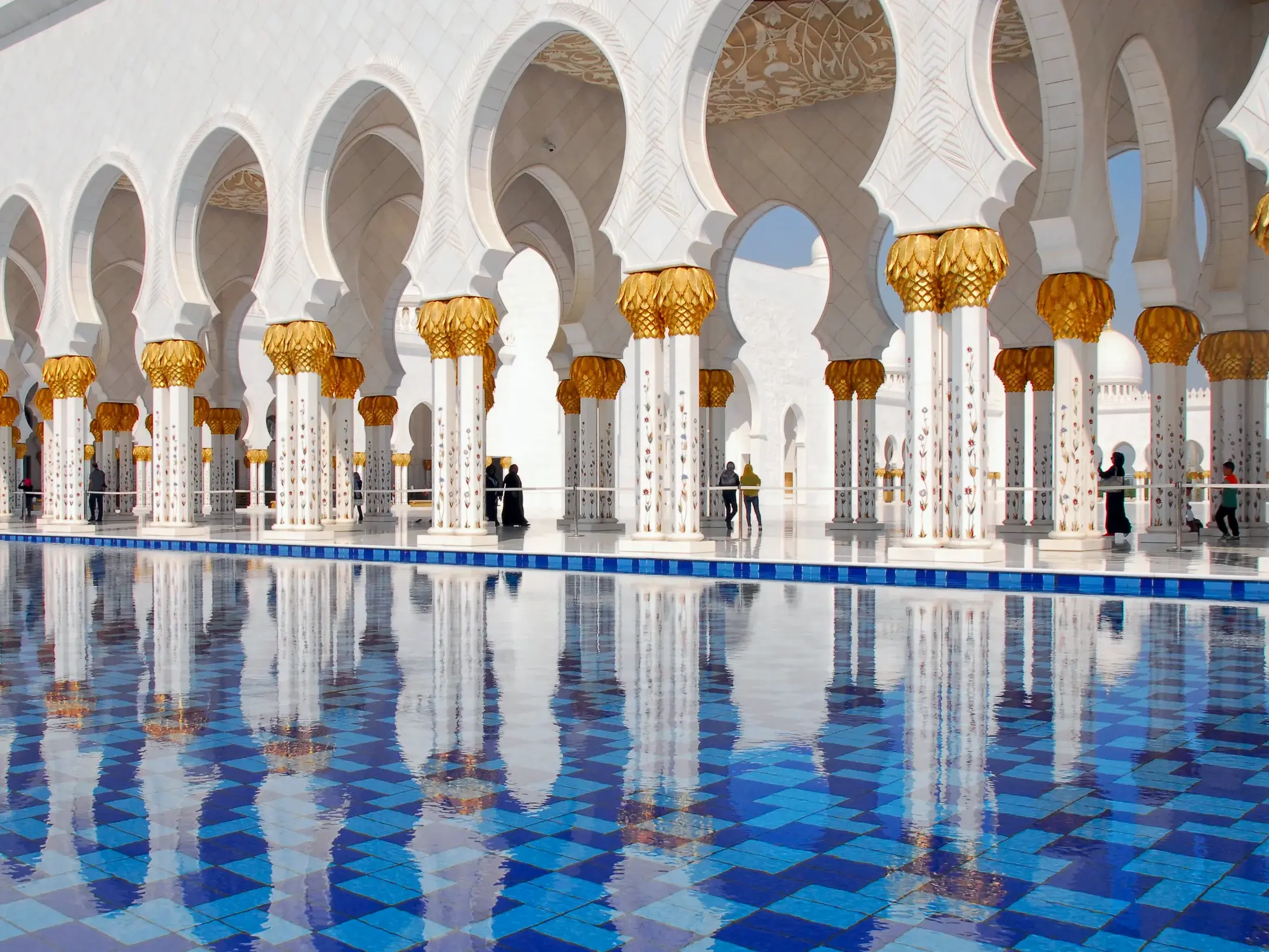 Weiße Marmorsäulen mit goldenen Kapitellen der Scheich-Zayed-Moschee in Abu Dhabi spiegeln sich in einem blau gekachelten Becken.