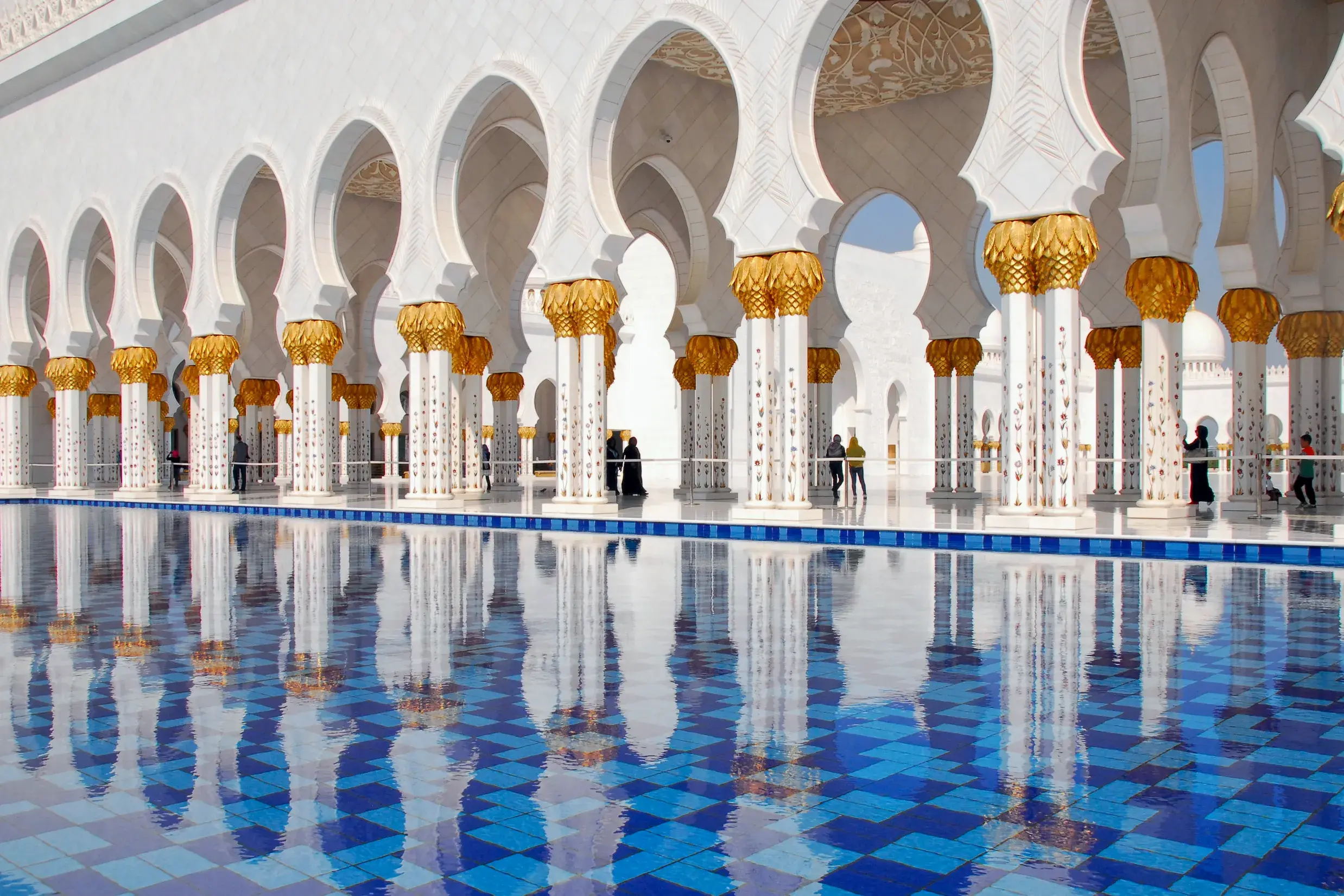 Weiße Marmorsäulen mit goldenen Kapitellen der Scheich-Zayed-Moschee in Abu Dhabi spiegeln sich in einem blau gekachelten Becken.