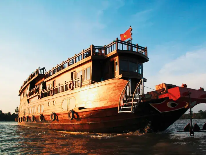 Ein traditionelles Holzschiff mit Vietnam-Flagge fährt während einer Kleingruppenreise bei Sonnenuntergang durch das Mekongdelta.