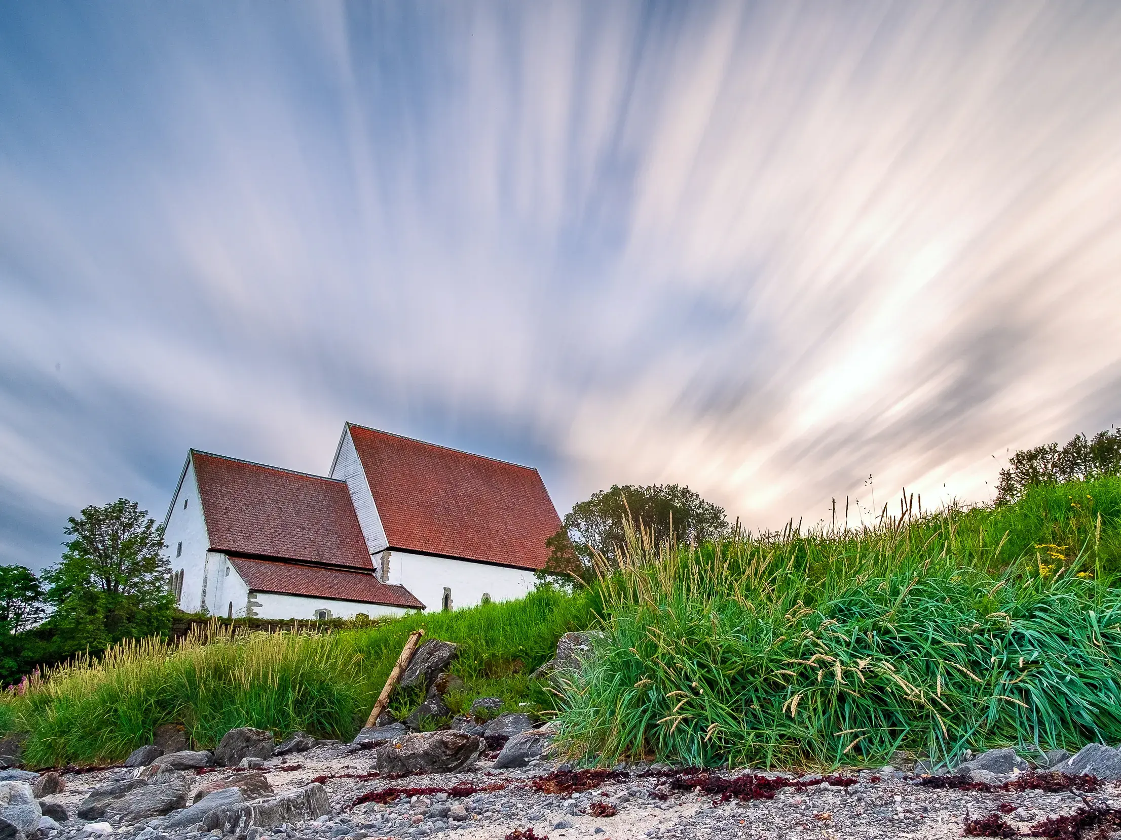 Die mittelalterliche Trondenes-Kirche in Norwegen auf einem grünen Hügel an der Küste unter einem Himmel mit Wolkenstreifen.