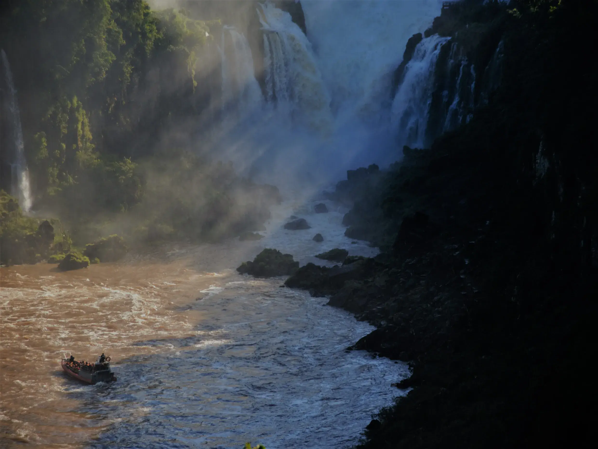 Eine Reisegruppe erlebt auf einer Bootsfahrt die Gischt der tosenden Wasserfälle an einer steilen Felswand in Brasilien.