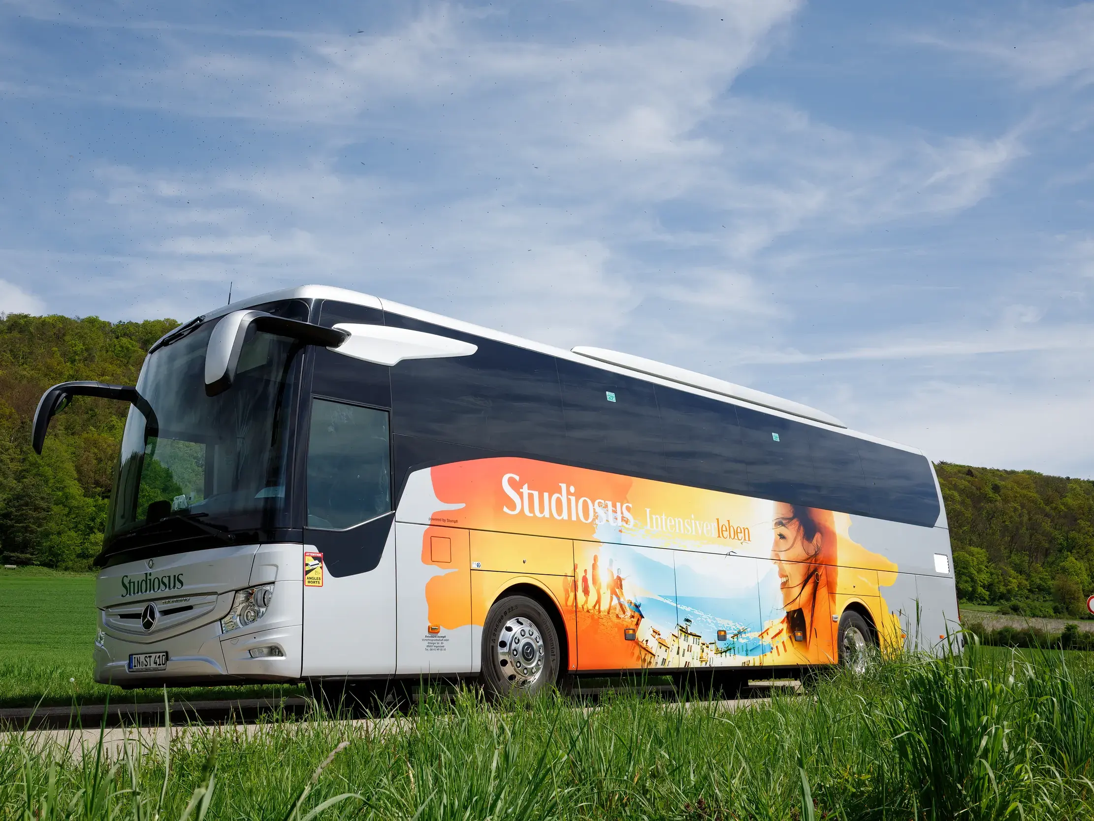Moderner Studiosus-Komfortbus parkt am Straßenrand vor einer grünen Wiese und einem bewaldeten Hügel unter blauem Himmel.