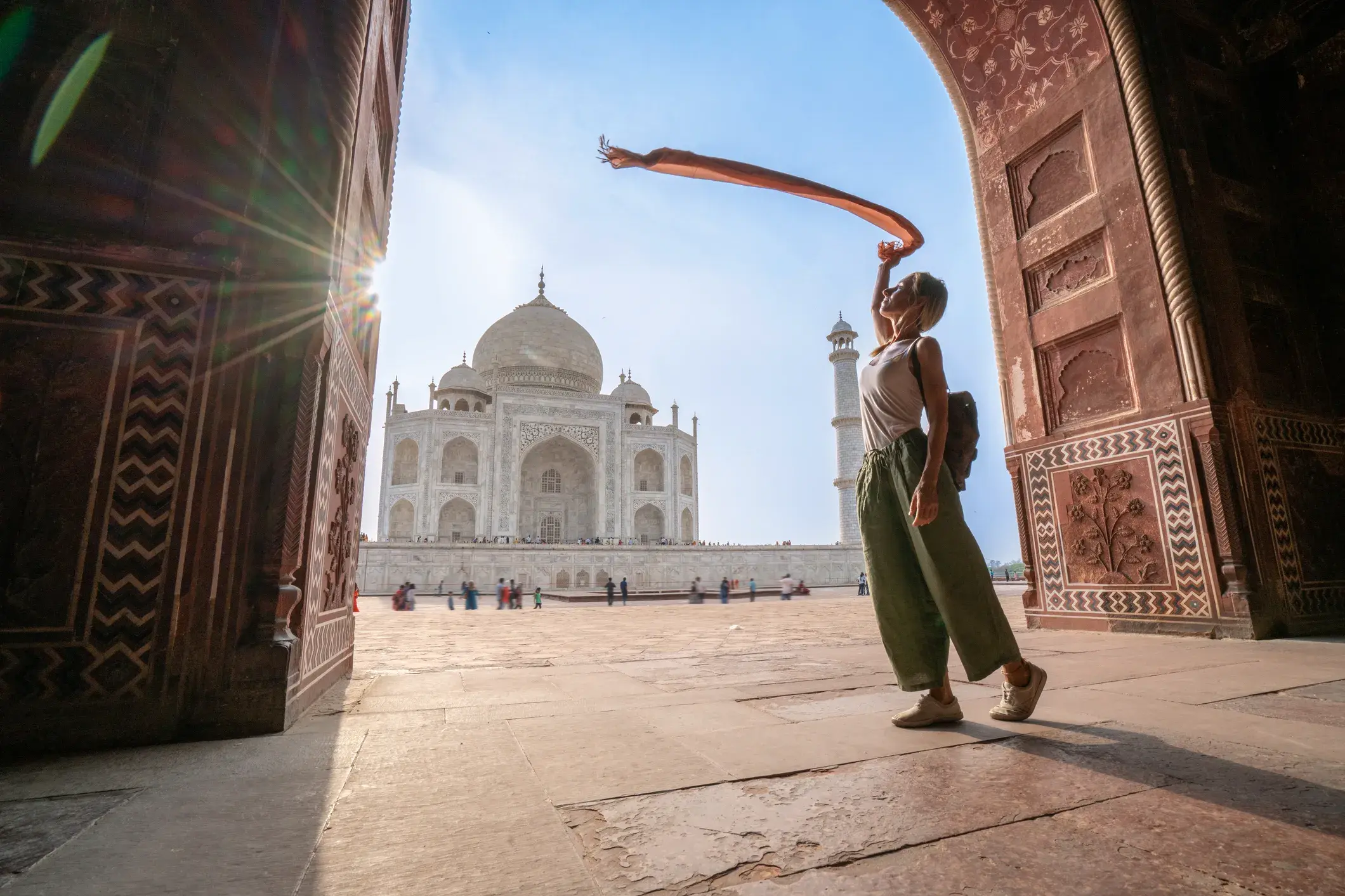 Junge Frau in einem verzierten Torbogen posiert mit einem wehenden Schal vor dem sonnenbeschienenen Taj Mahal in Agra.
