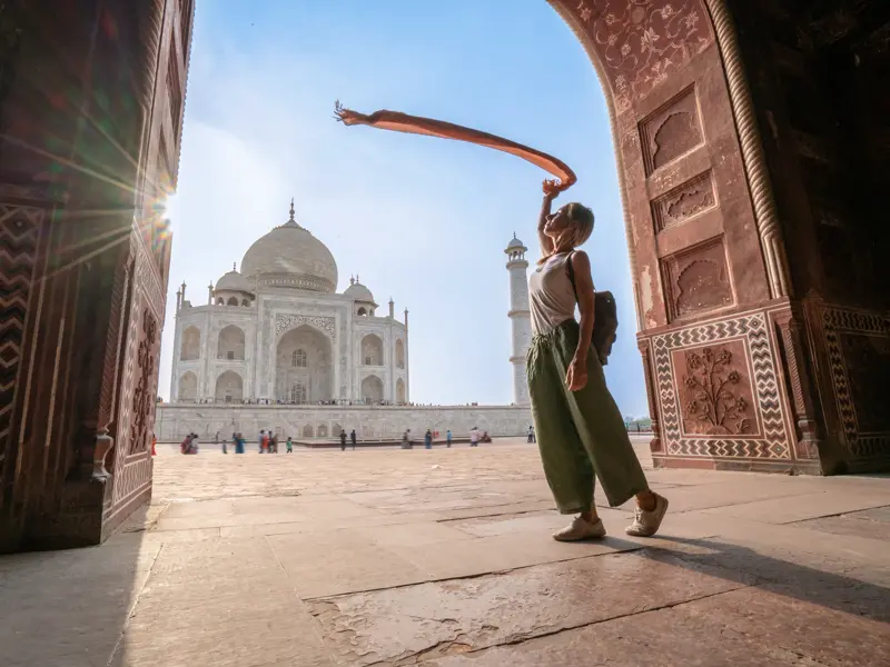 Junge Frau in einem verzierten Torbogen posiert mit einem wehenden Schal vor dem sonnenbeschienenen Taj Mahal in Agra.