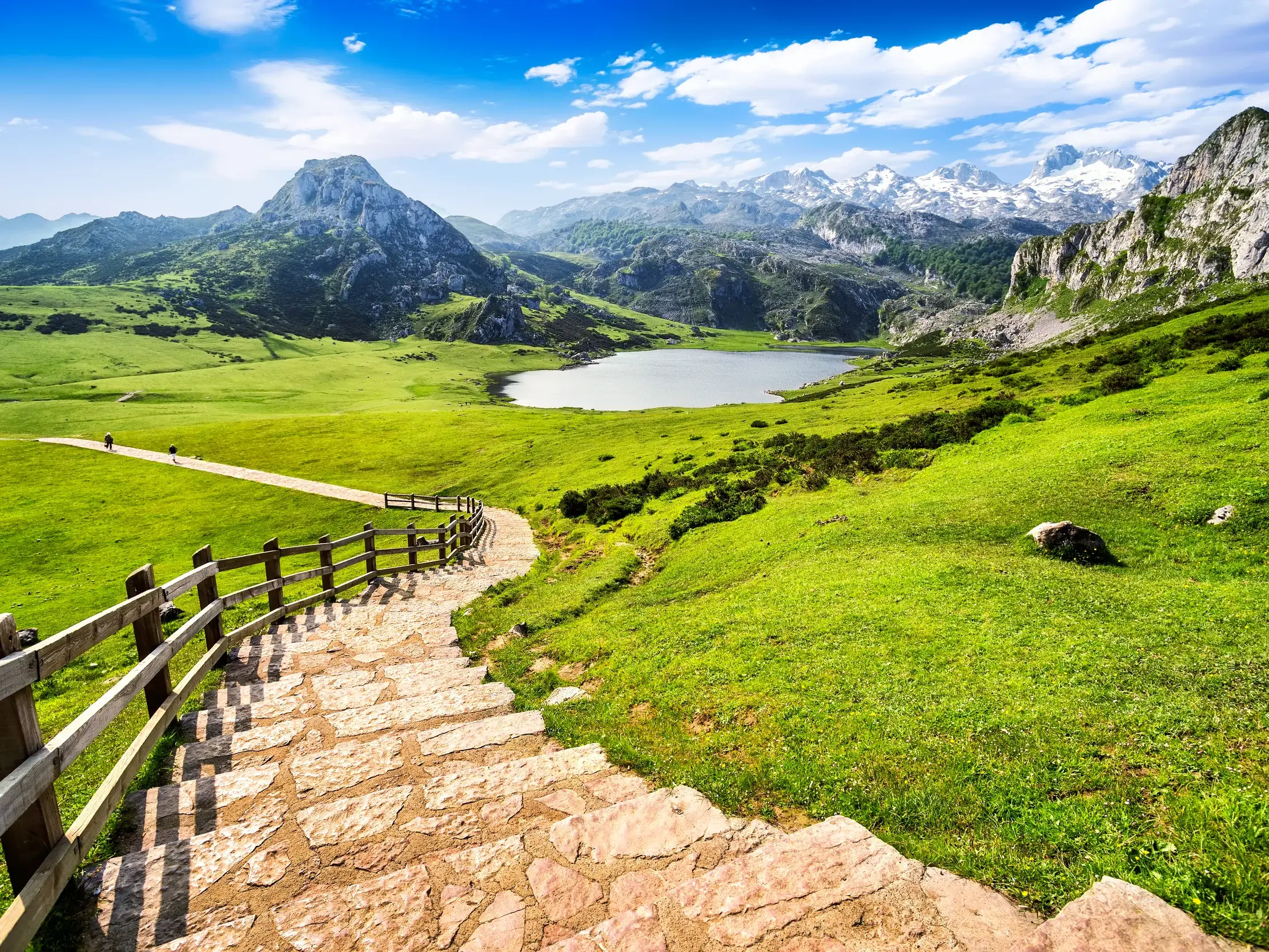 Eine Steintreppe führt hinab in die weite, grüne Landschaft des Nationalparks Picos de Europa mit Bergsee und den schroffen, noch mit Schnee bedeckten Bergen.